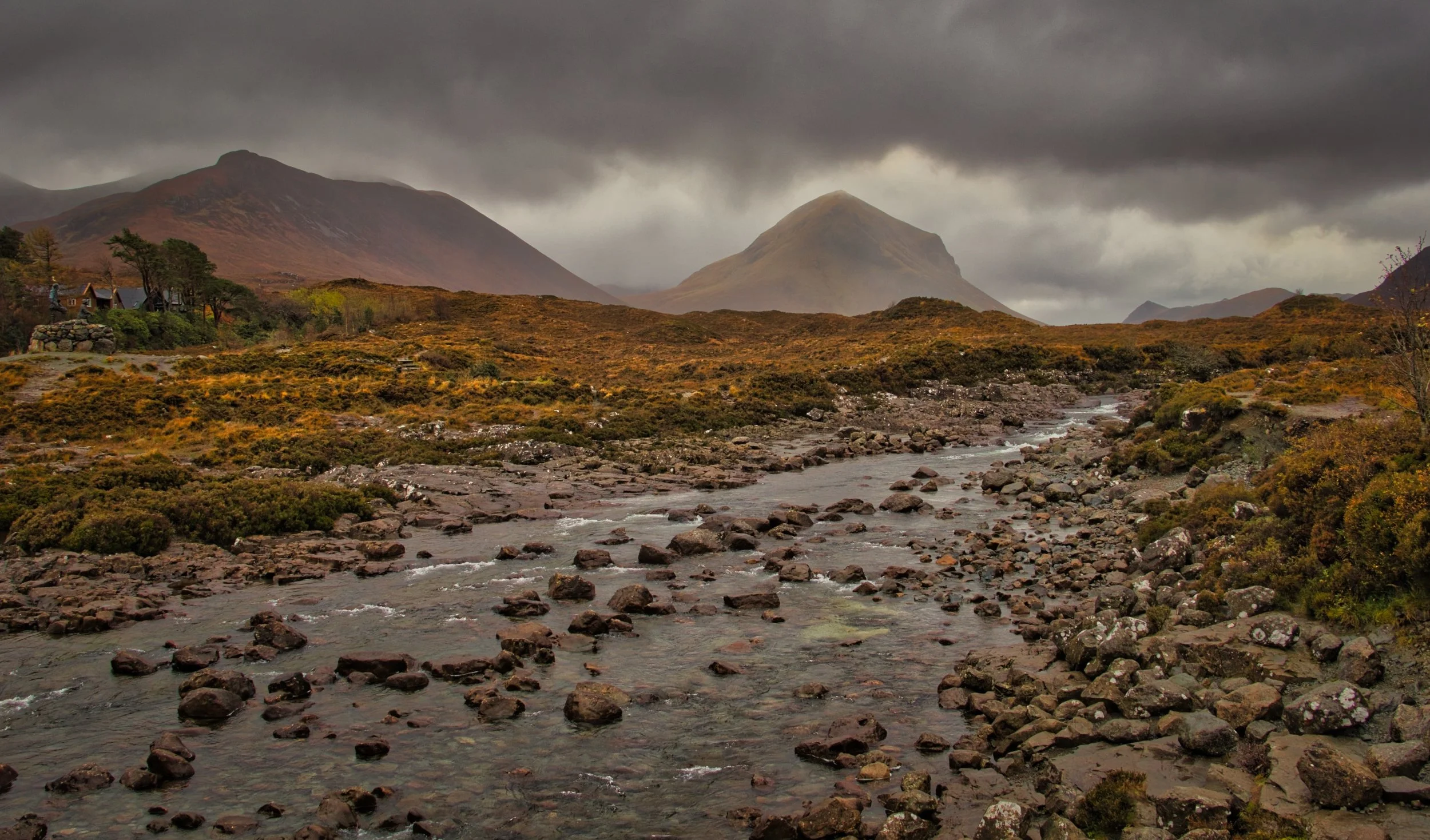 The wide, rocky bed of the River Sligachan on the Isle of Skye, winding through rich copper moorland beneath the dramatic peaks of the Red Cuillins under heavy storm clouds. Evocative fine art Scottish landscape photography capturing the moody atmosp