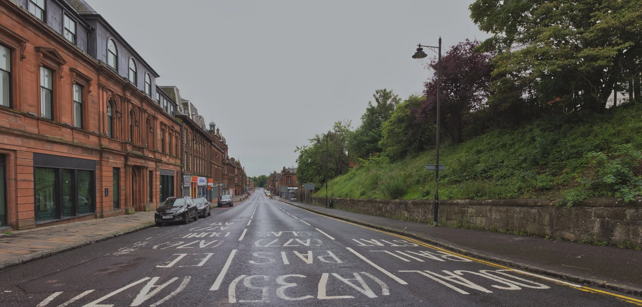 The wide, empty tarmac of John Finnie Street in Kilmarnock, showing painted road markings (A735, Paisley, A77) leading past grand red sandstone architecture under a flat grey sky. Deadpan urban landscape photography capturing perspective and quiet to