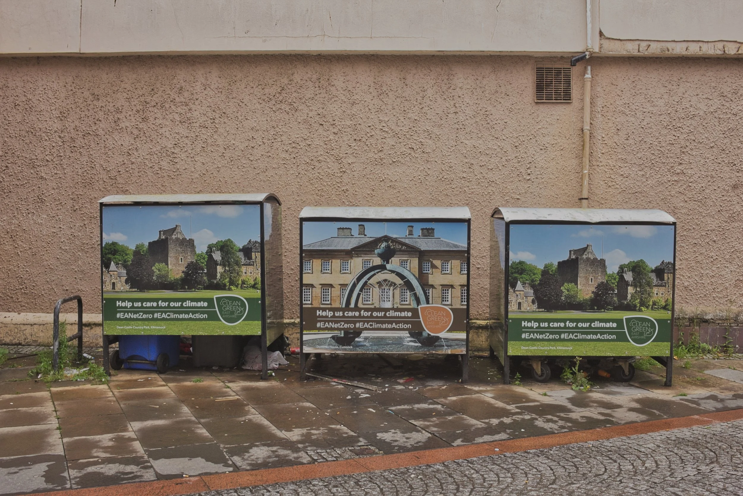 Three public waste bins wrapped in marketing imagery of local historic estates, positioned against a textured pink pebbledash wall on wet pavement in East Ayrshire. Deadpan urban landscape photography capturing the quiet humour and contrast of modern