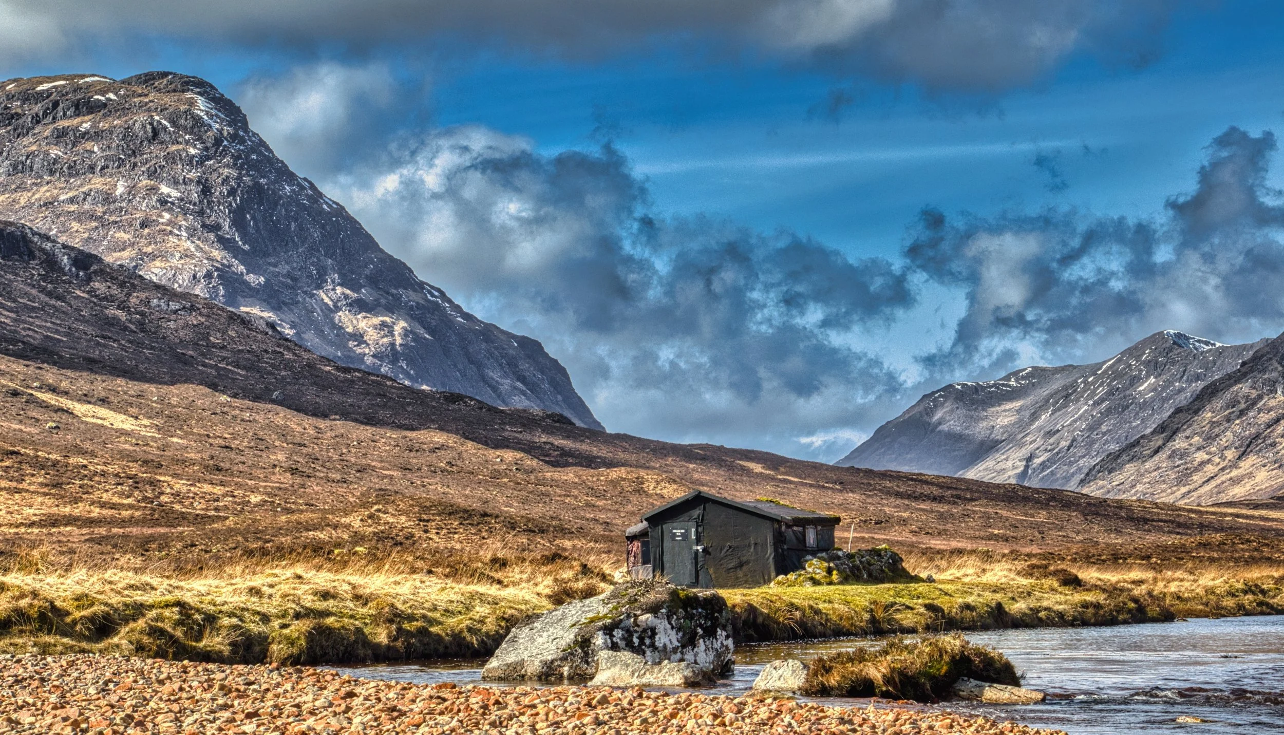 The iconic black climbing hut nestled on the rugged valley floor of Glencoe, flanked by towering Highland peaks under a dramatic, cloud-swept sky. Evocative Scottish landscape photography capturing the isolation, golden moorland, and wild scale of th