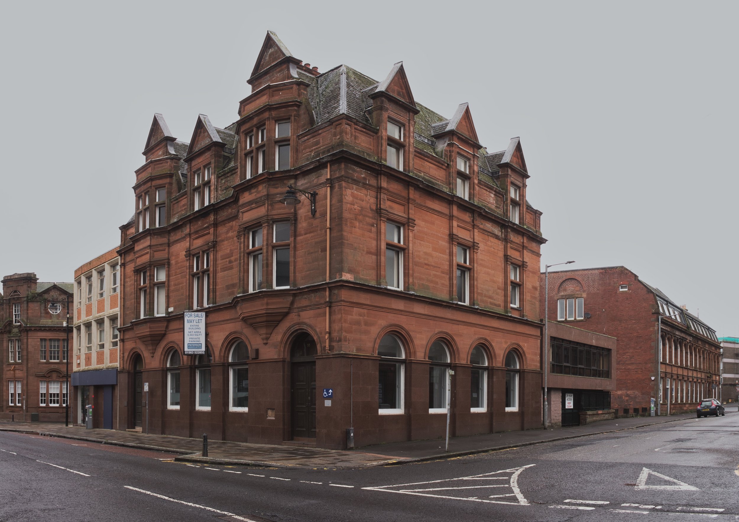 An imposing red sandstone corner building with arched windows and a commercial 'For Sale' sign under a flat grey sky in East Ayrshire. Deadpan urban landscape photography capturing the quiet geometry of heritage architecture and high street vacancies