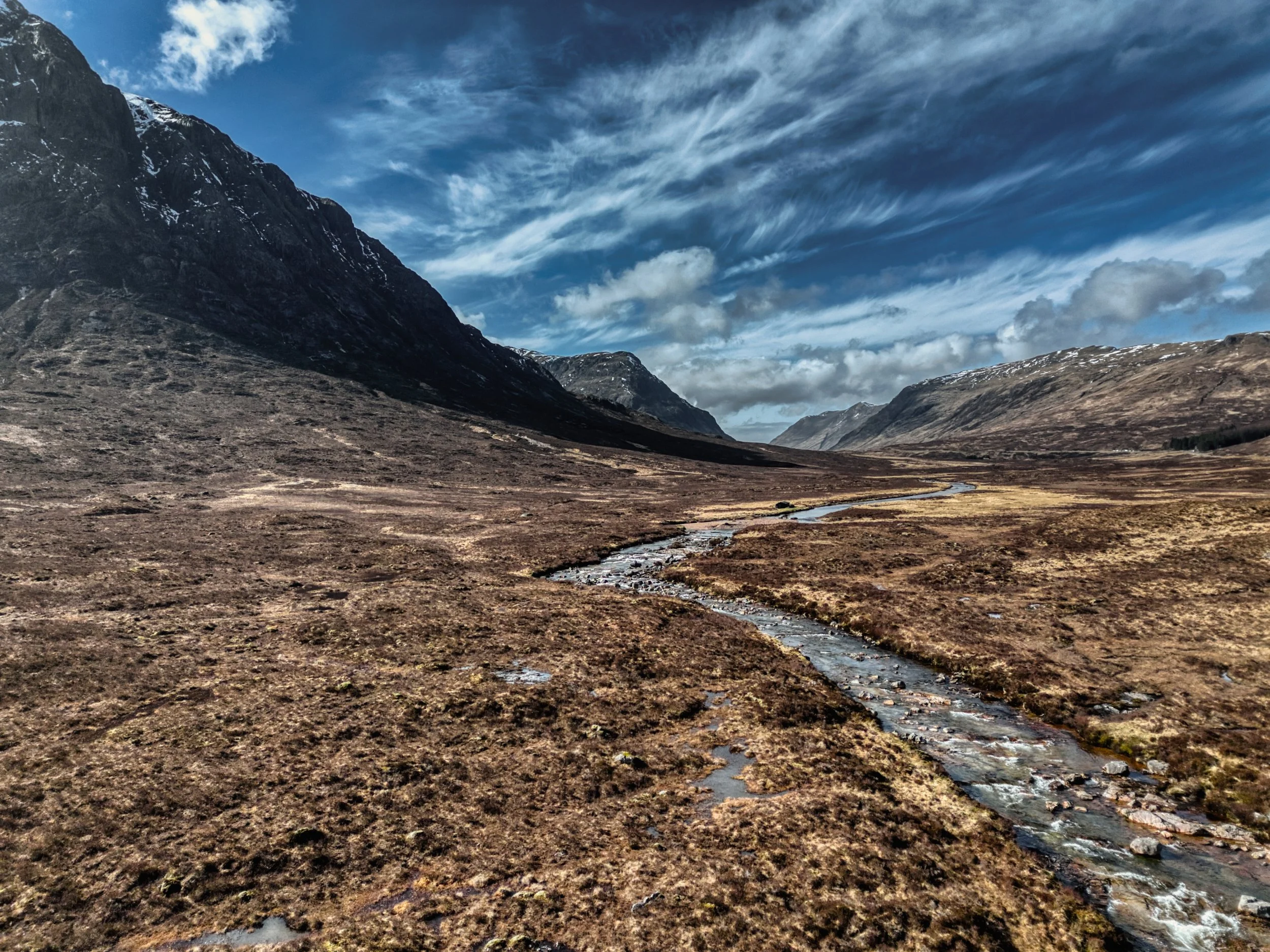 An aerial drone photograph looking down a vast mountain glen, with the winding River Coupall snaking through the golden, rugged moorland under a dramatic sky. Fine art Scottish landscape photography capturing the immense scale of the Highlands from a