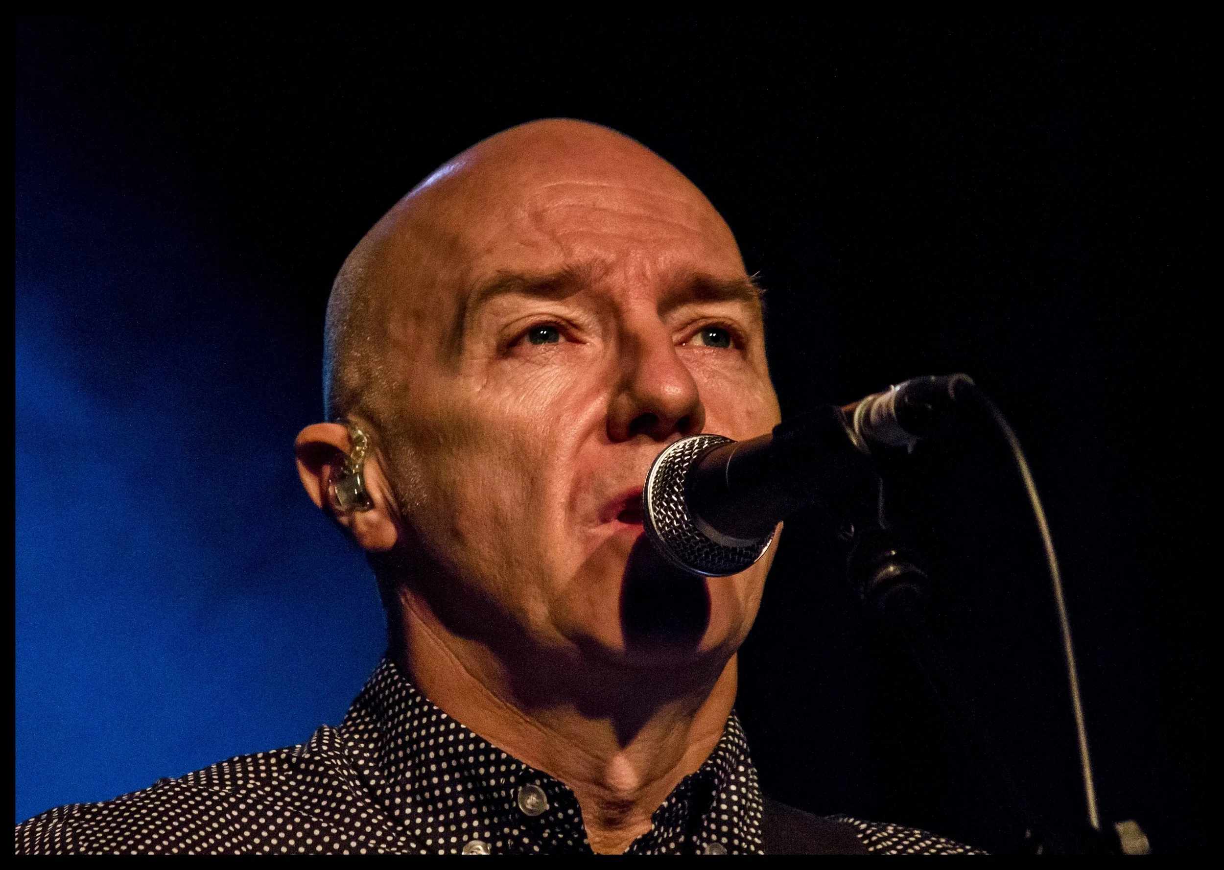 A close-up live concert portrait of singer Midge Ure performing at a microphone under dramatic blue stage lighting. Intimate music photography capturing the intense focus and expression of a classic electronic synth-pop and rock artist.