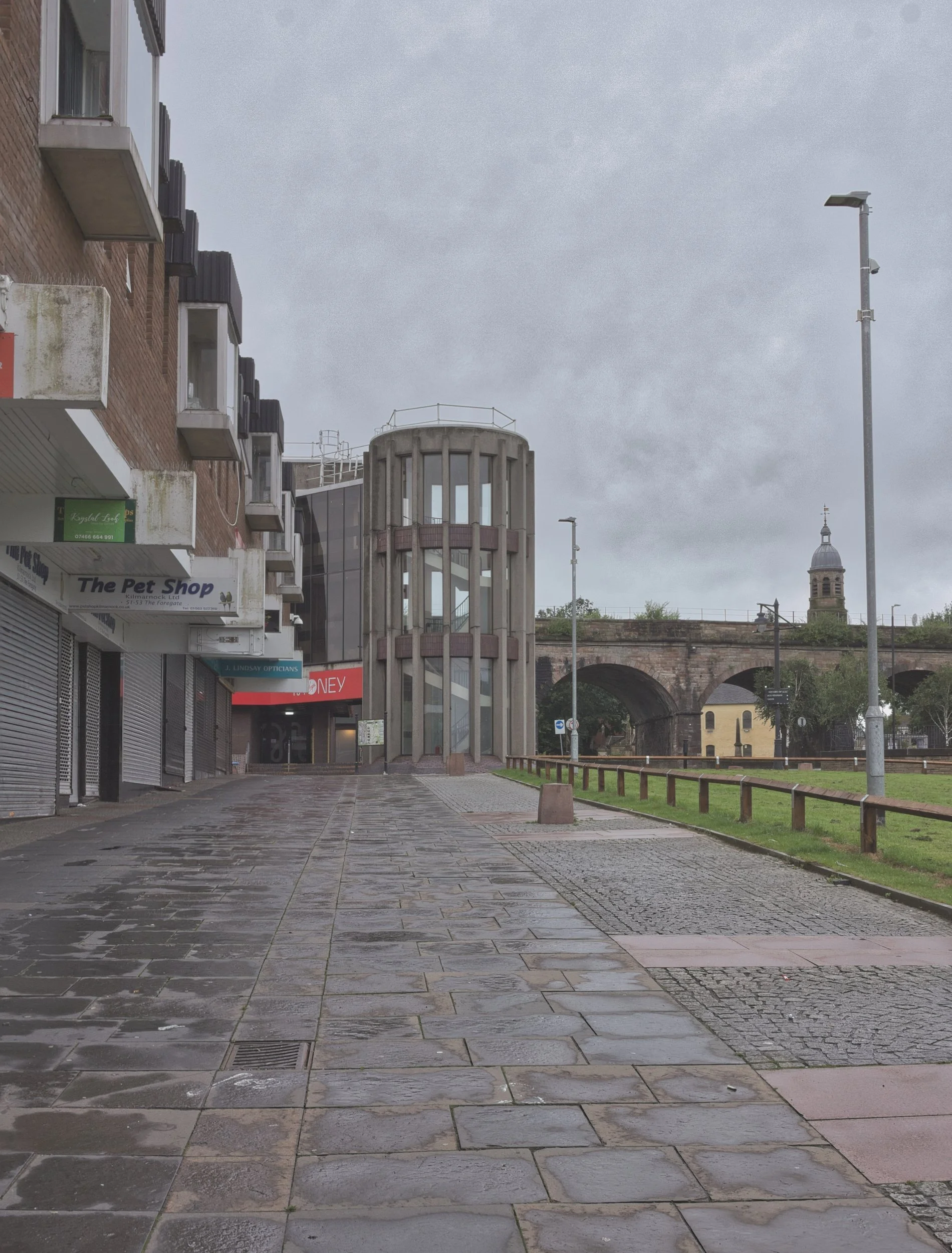 A damp pedestrian precinct leading towards a brutalist concrete stairwell tower, flanked by shuttered shops and a historic stone railway viaduct under a flat grey sky in Kilmarnock, East Ayrshire. Deadpan urban landscape photography capturing the qui