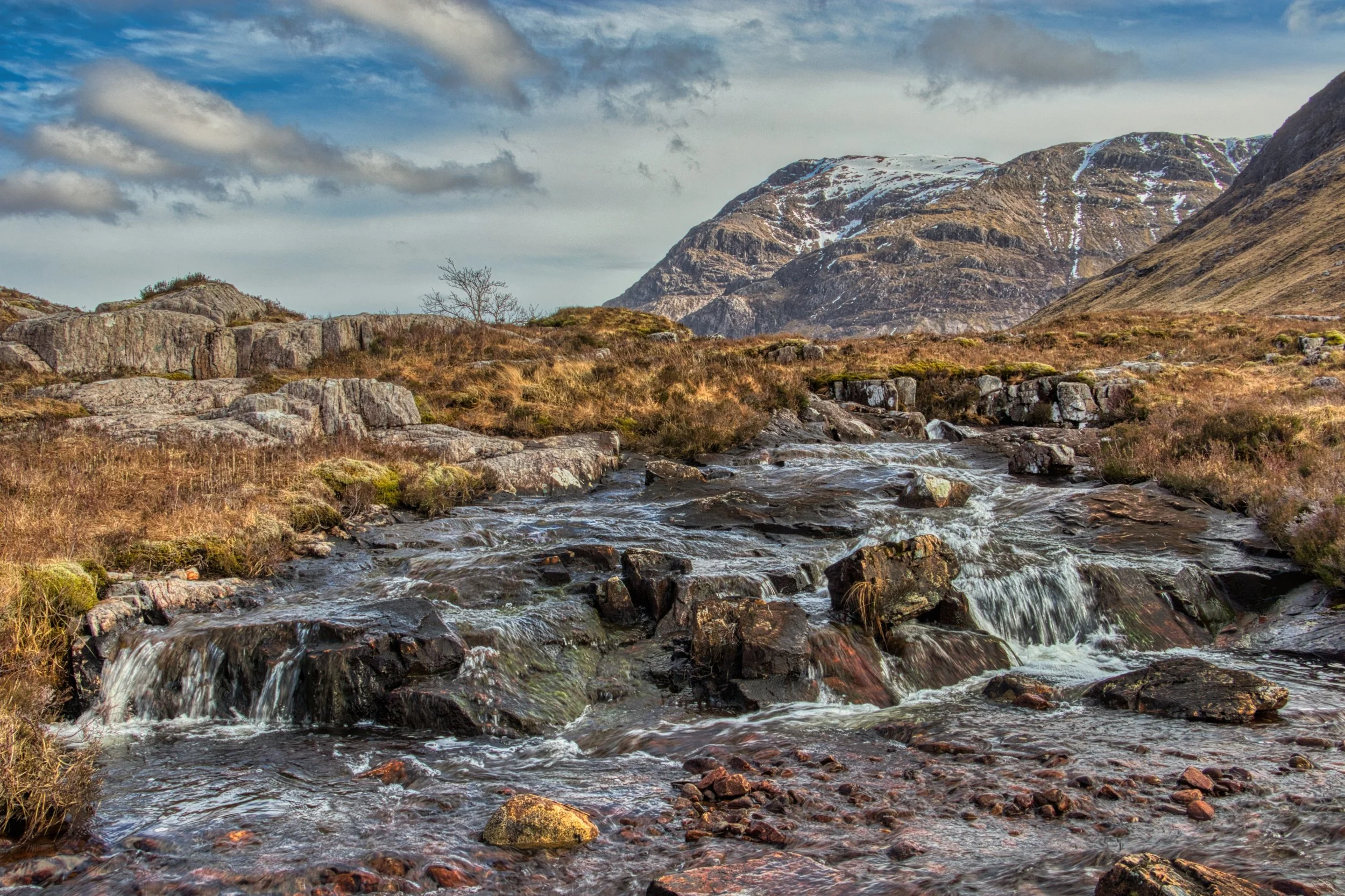 A rocky mountain stream flowing over dark stones in Glencoe, with a textured Highland peak rising under a dramatic, cloud-filled sky. Atmospheric Scottish landscape photography capturing the rugged textures and wild expanse of the glen.