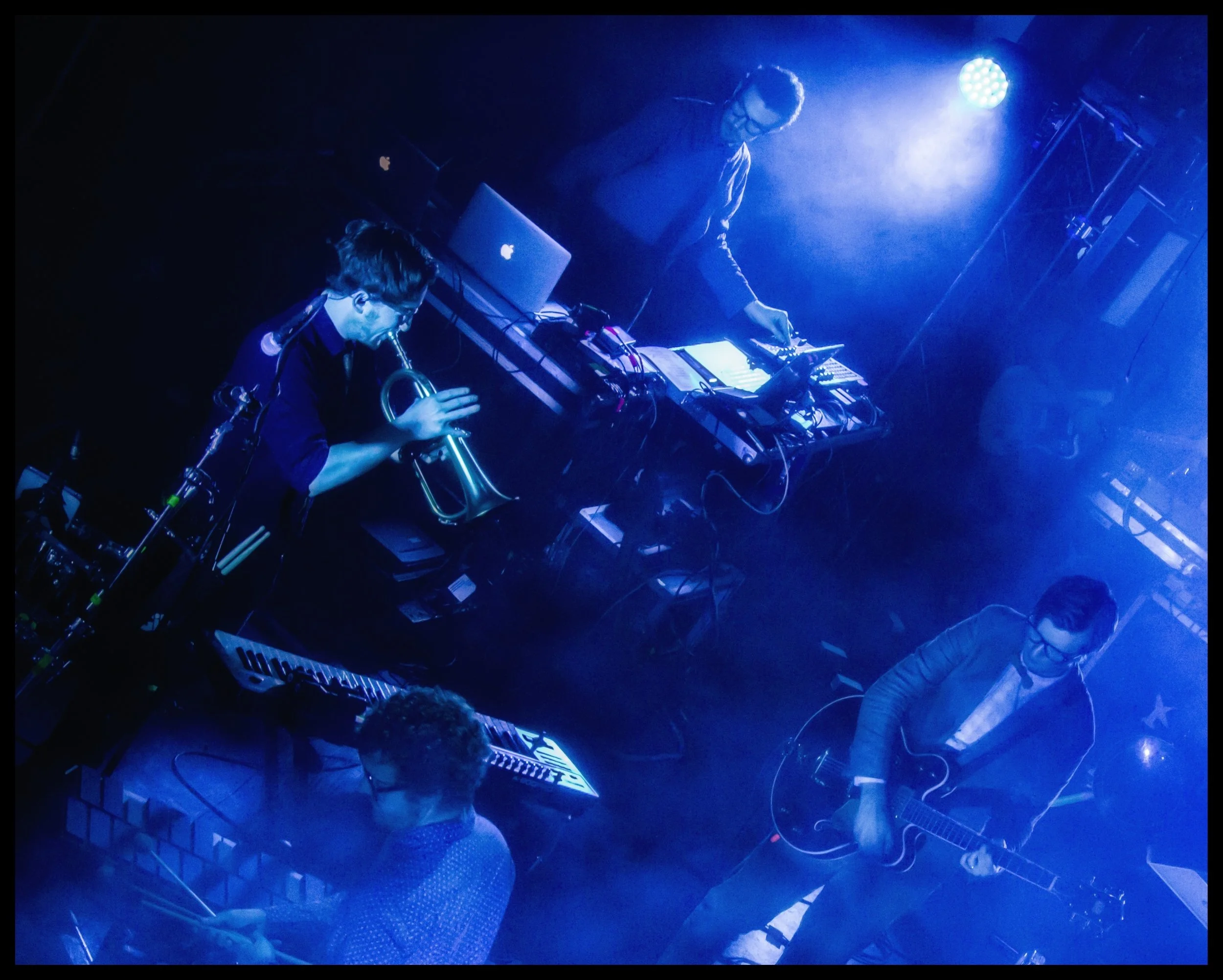 An overhead live concert photograph of the band Public Service Broadcasting performing on a smoke-filled stage under intense blue spotlights. Dynamic live music photography capturing a trumpet player, guitarist, and electronic synthesizer setups from