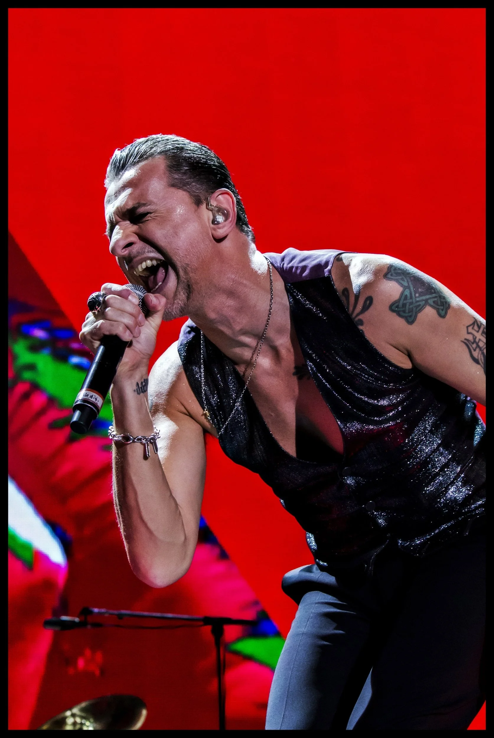 Depeche Mode frontman Dave Gahan singing passionately into a microphone on a vibrant red stage backdrop during a live concert performance. Dynamic music photography capturing the high-energy showmanship and grit of an electronic rock gig.