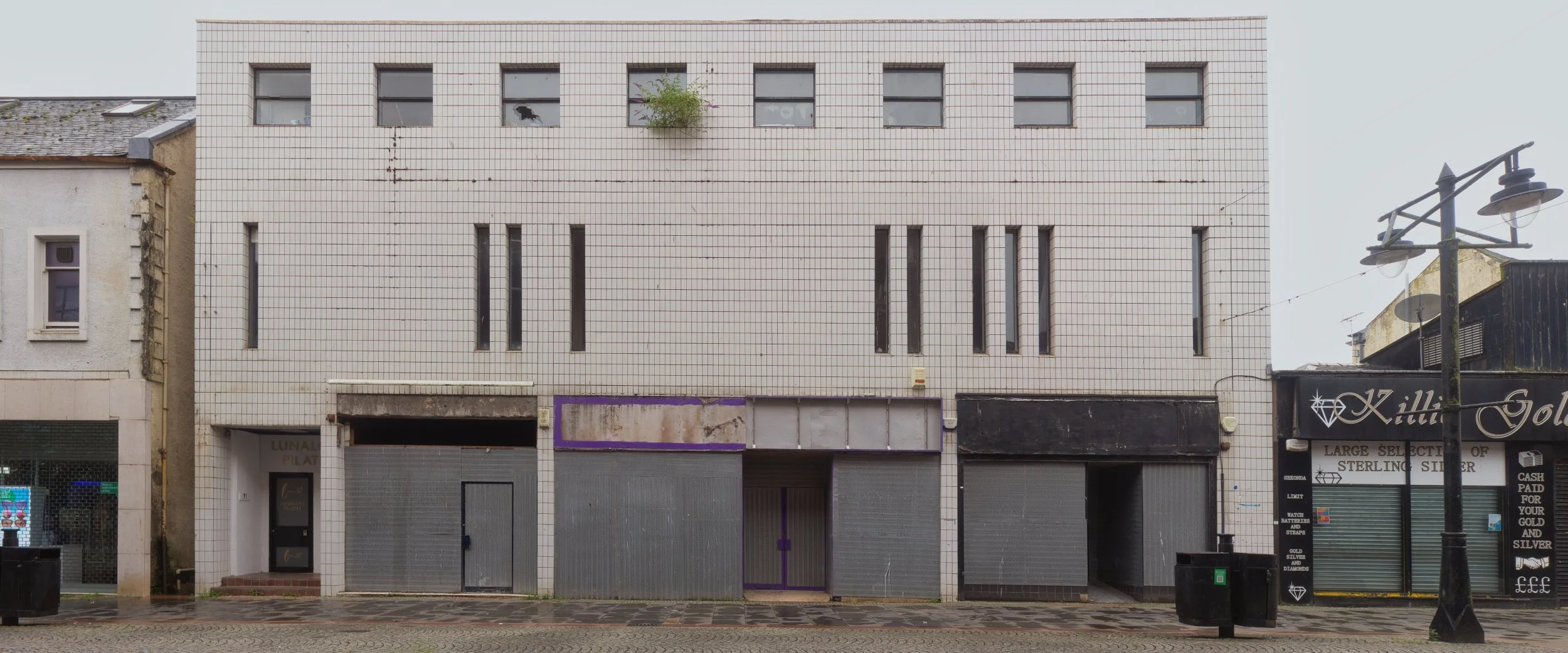 A wide, multi-fronted commercial building facade covered in white square tiles with closed metal security shutters under an overcast sky in Kilmarnock, East Ayrshire. Deadpan urban landscape photography capturing modern high street architecture and t