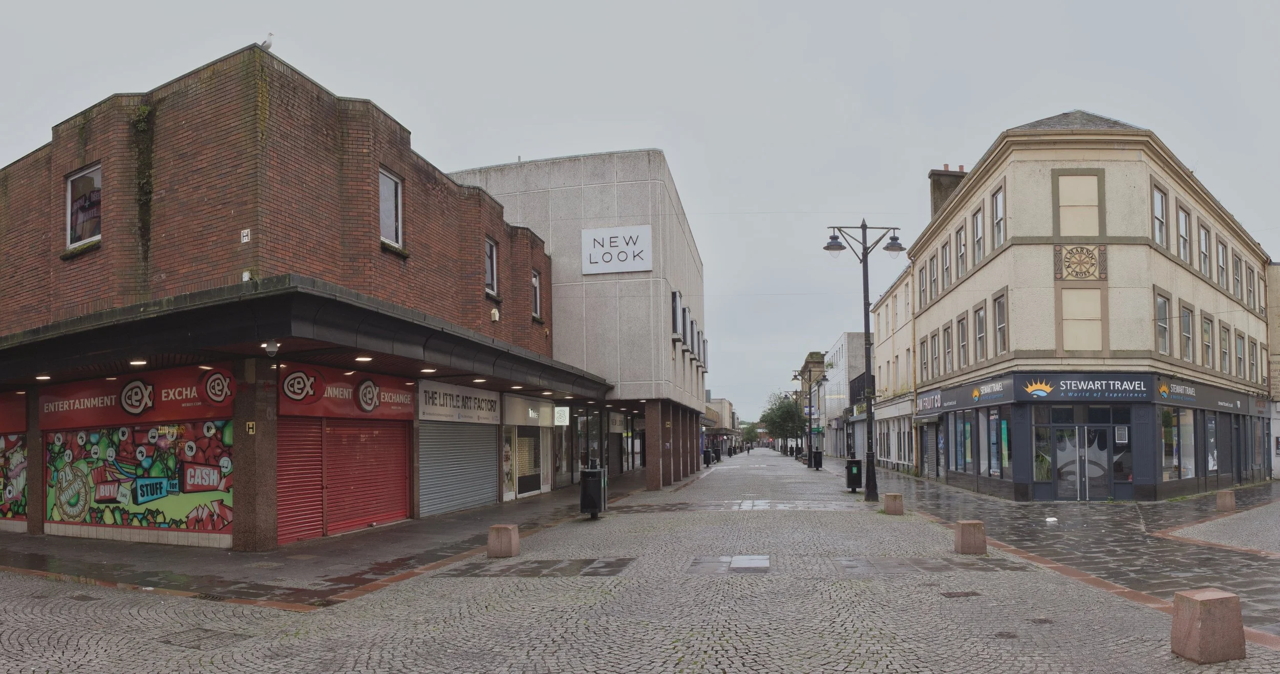 A wide pedestrianised high street lined with modern retail storefronts, shuttered commercial units, and traditional corner buildings under a flat grey sky in Kilmarnock, East Ayrshire. Deadpan urban landscape photography capturing the quiet perspecti