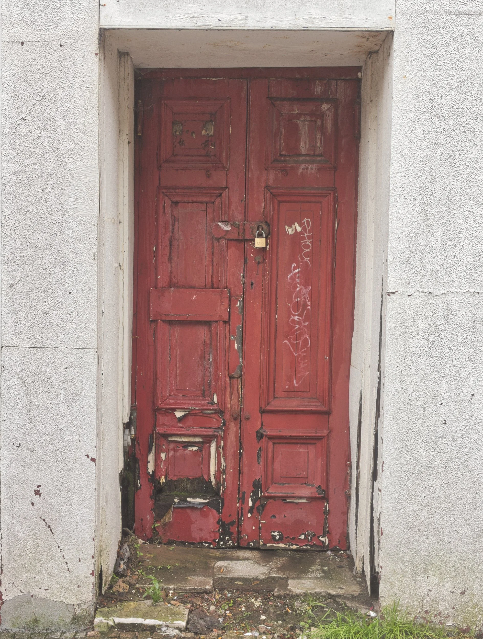 A weathered, peeling red wooden double door padlocked shut and set into a cracked white rendered wall in East Ayrshire. Deadpan urban landscape photography capturing the stark textures and quiet details of town decay.