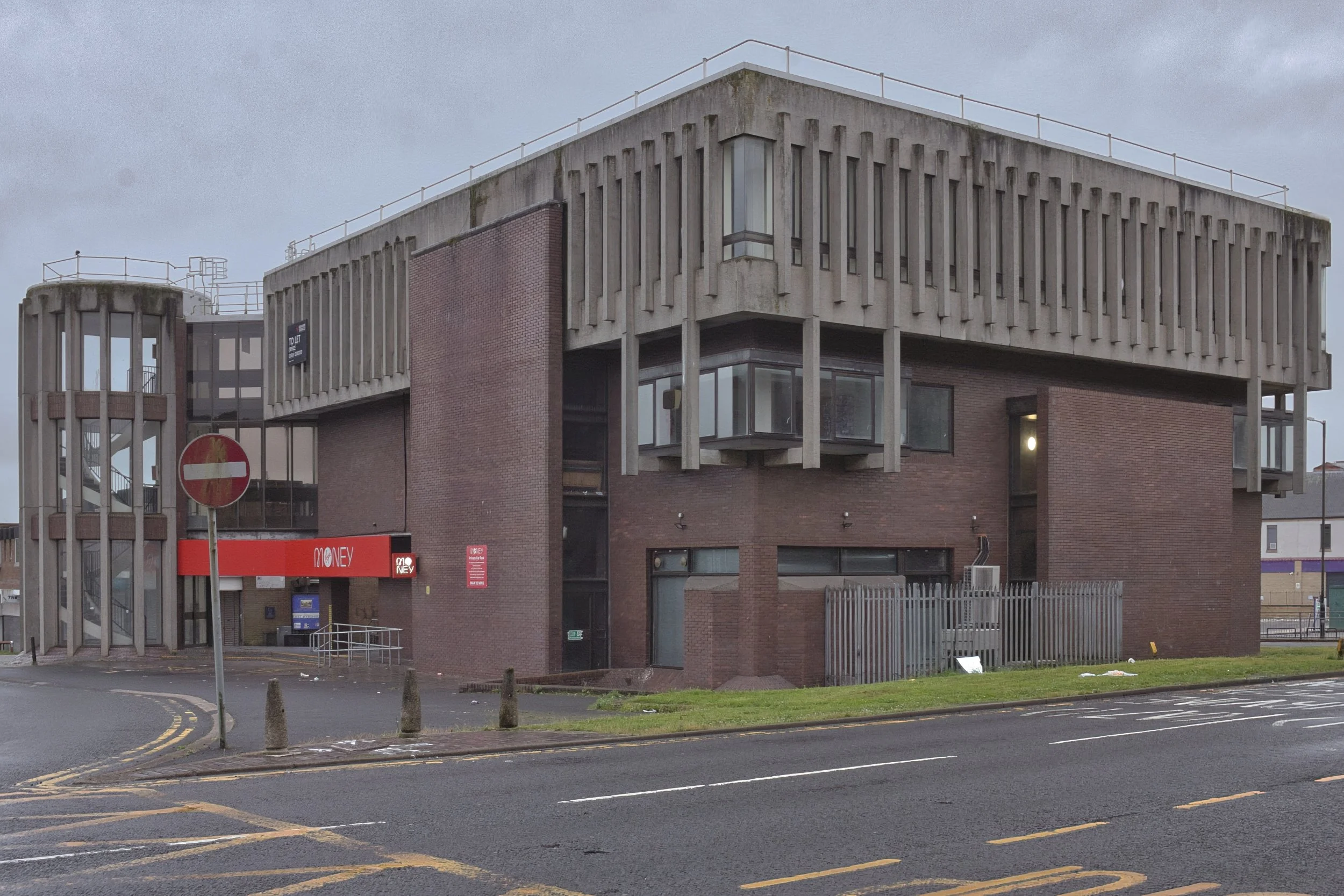 A brutalist concrete and dark brick commercial building featuring a prominent bright red 'MONEY' sign and a faded No Entry road sign on a damp street corner under an overcast sky in Kilmarnock, East Ayrshire. Deadpan urban architecture photography ca