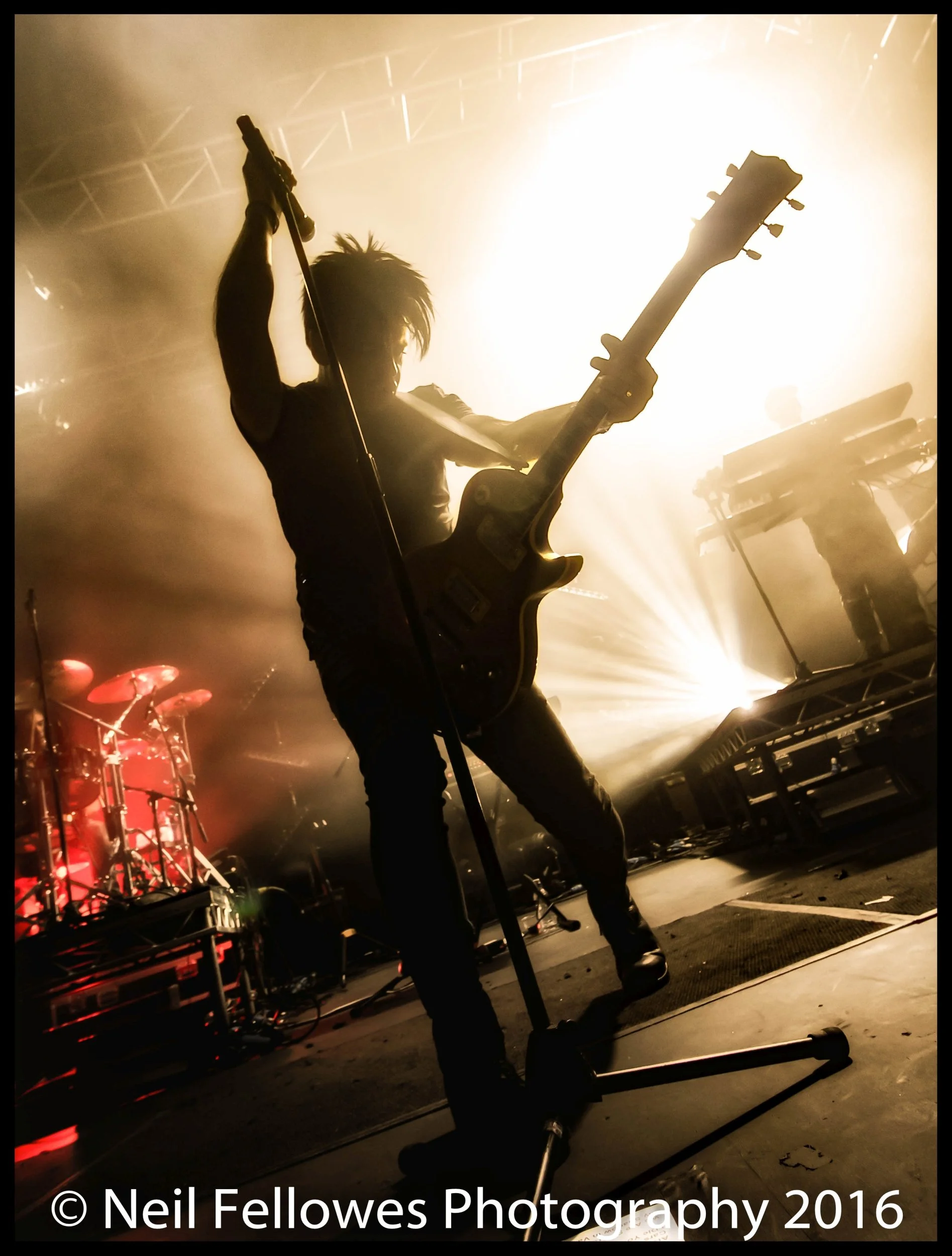 A dramatic live concert silhouette of electronic music pioneer Gary Numan performing on a smoke-filled stage, backlit by an intense white spotlight with sharp light rays piercing the darkness. High-contrast concert photography capturing the raw energ
