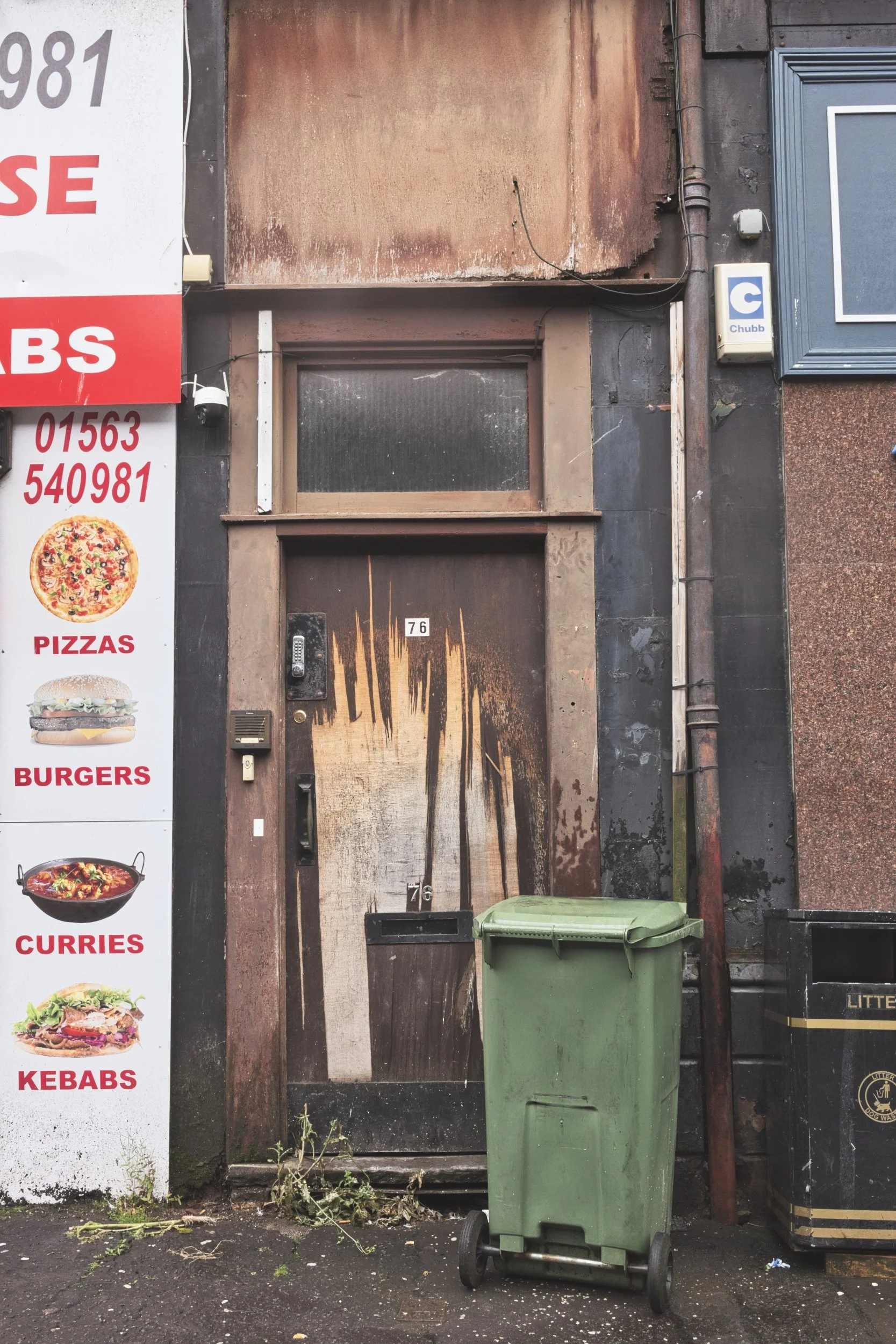 A heavily weathered and scratched wooden door numbered 76, fronted by a green wheelie bin and flanked by a fast-food takeaway sign in Kilmarnock, East Ayrshire. Deadpan urban architecture photography capturing the textures of high street decay.