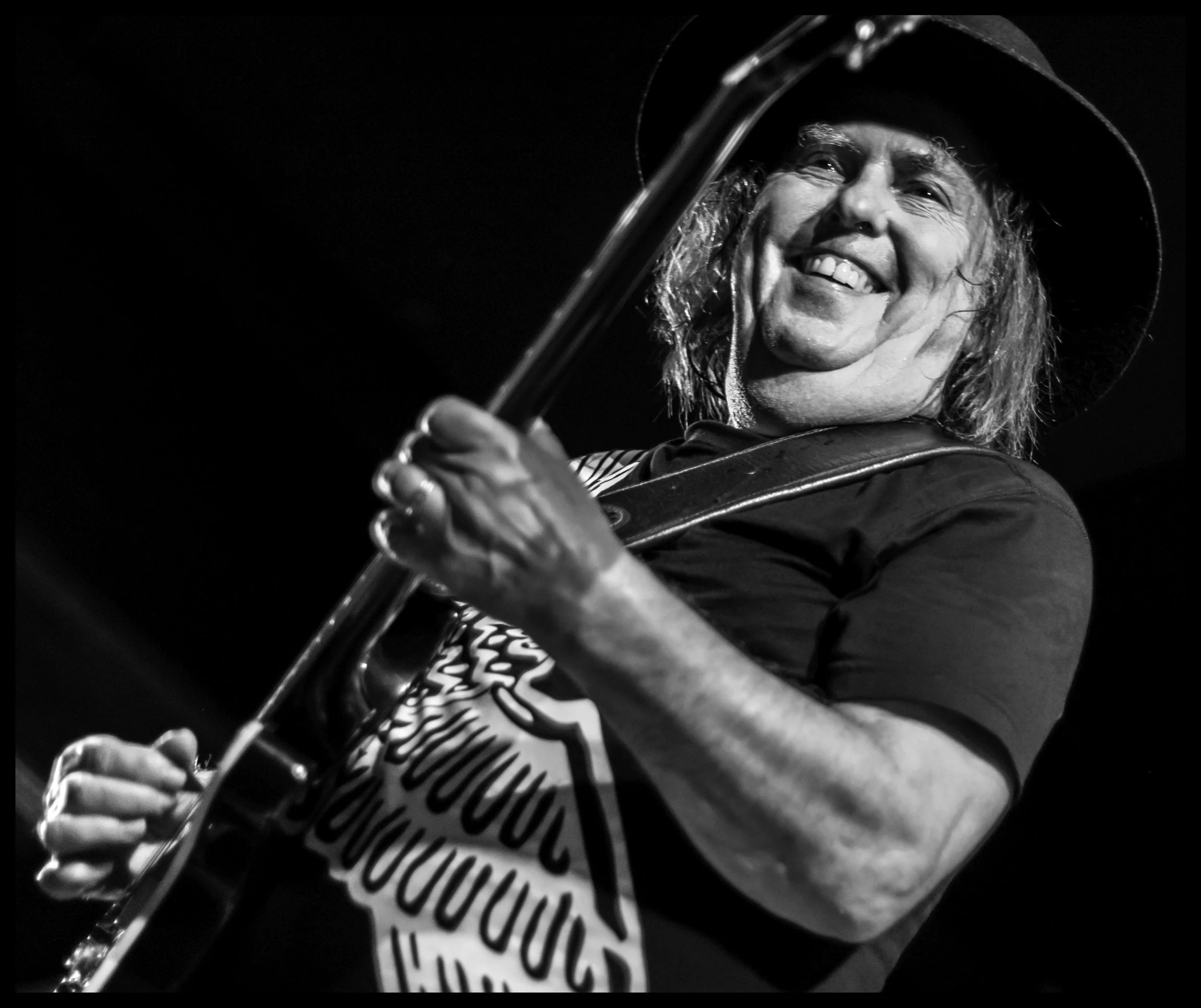 A high-contrast black and white concert photograph of Slade guitarist Dave Hill smiling warmly while performing on stage with his guitar. Expressive live music photography capturing the iconic energy and performance details of classic British glam ro
