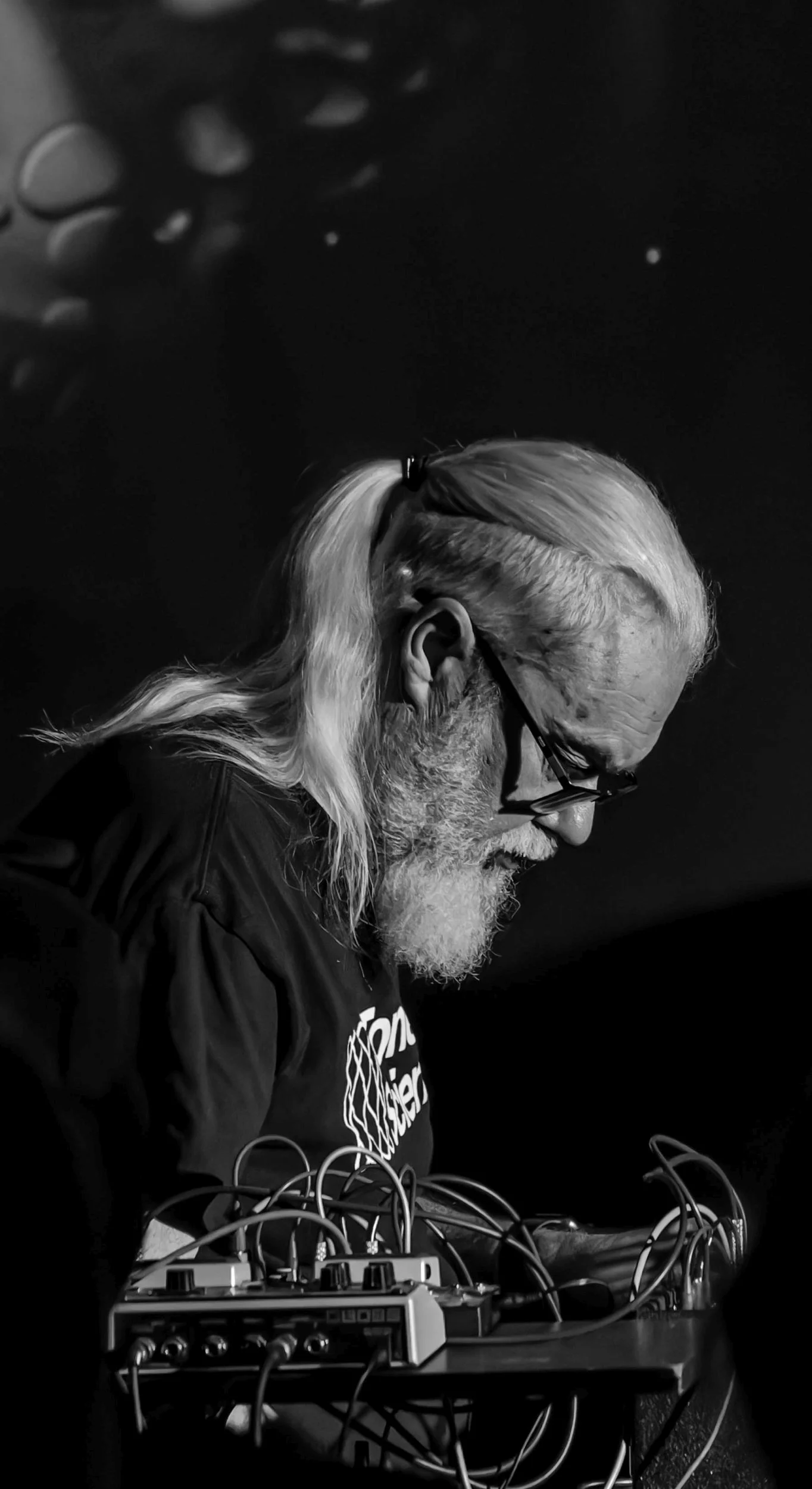 An atmospheric black and white concert portrait of electronic ambient musician Ian Boddy in profile, operating a synthesizer setup with patch cables on a dark stage. Specialised live music photography capturing analogue synth performance and creative