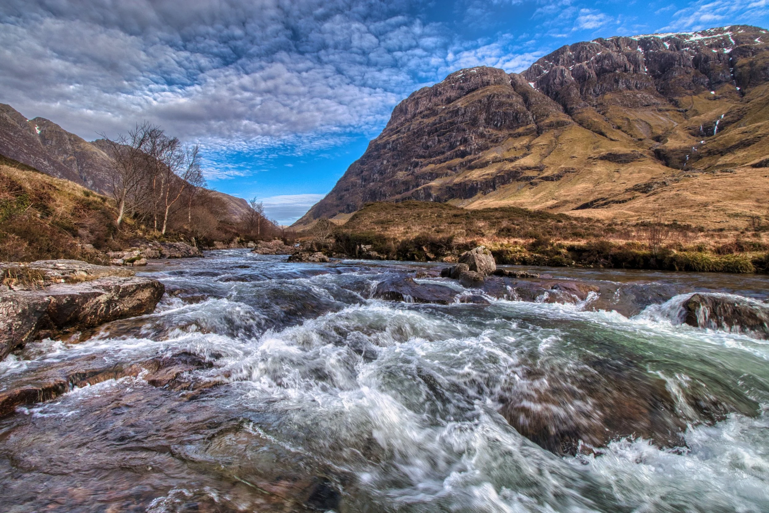 The rushing, white waters of the River Coe carving through a dramatic Highland glen, framed by towering, rugged mountain peaks under a patterned sky. Atmospheric Scottish landscape photography capturing the movement, texture, and grand scale of Glenc