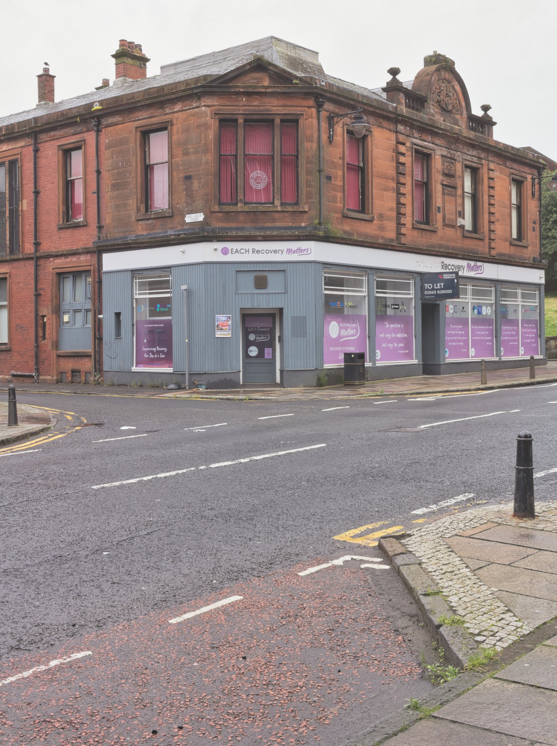 A historic red sandstone corner building featuring a modern blue and purple recovery service storefront with a 'To Let' sign on a wet street in Kilmarnock, East Ayrshire. Deadpan urban landscape photography documenting architectural layers and high s