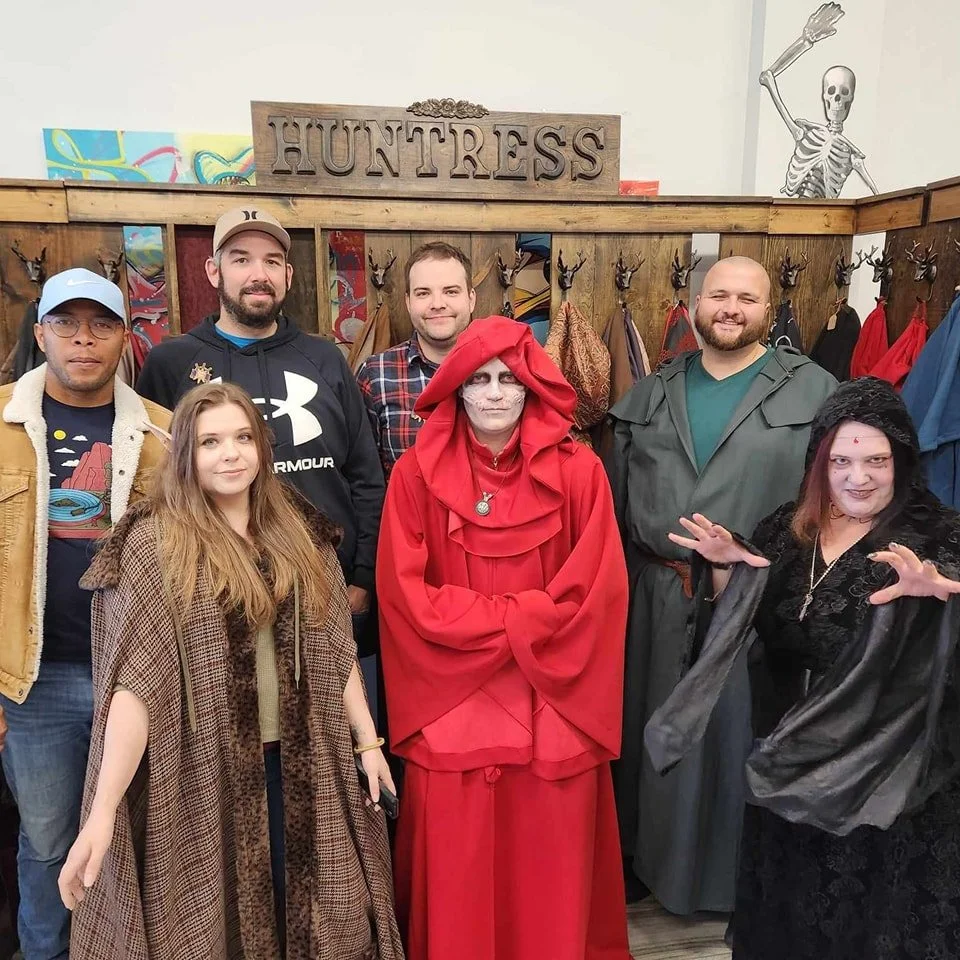 Group of seven people dressed in Halloween costumes at a store called 'Huntress,' with a skeleton decoration in the background.