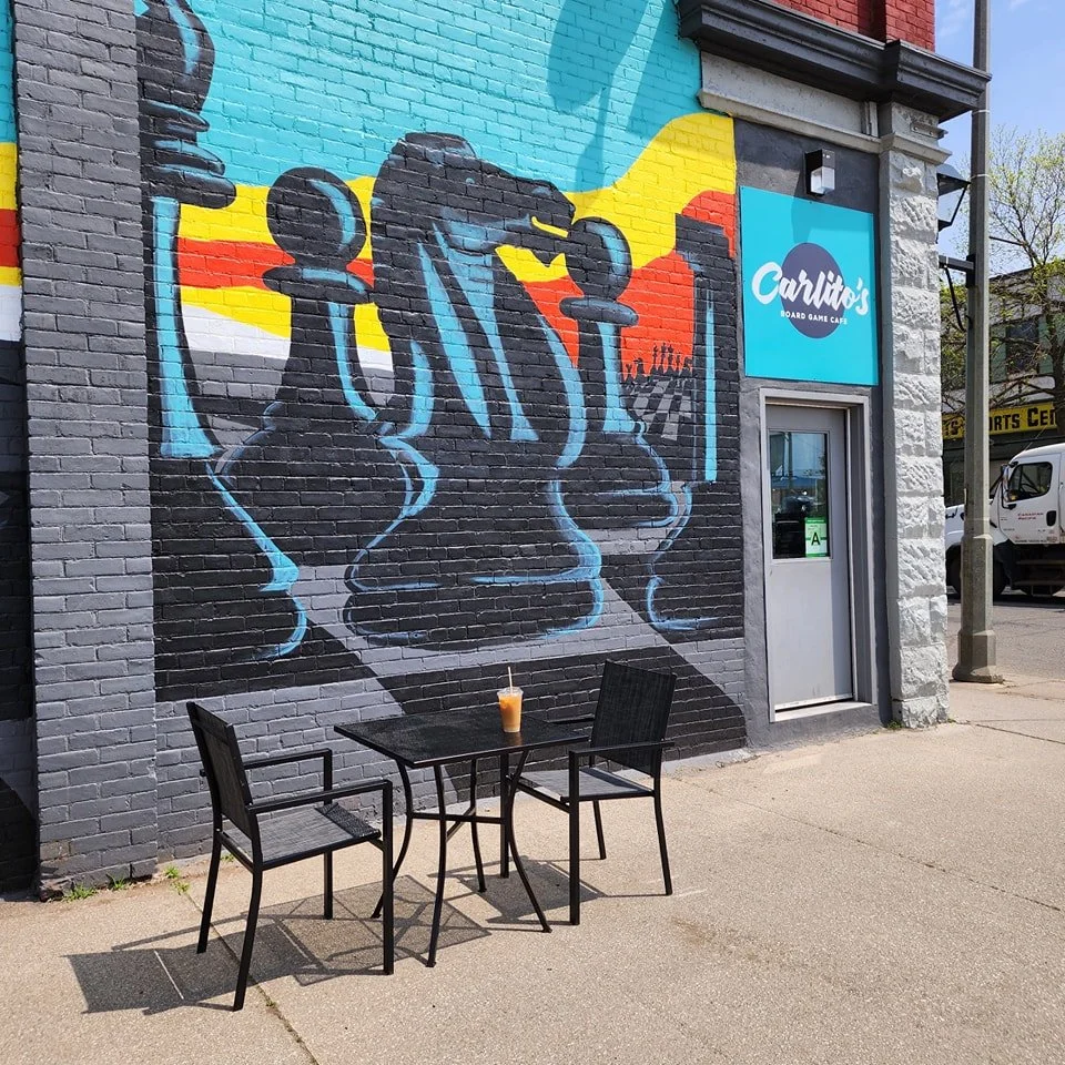 Street scene outside Carlito's Board Game Cafe with a large mural of chess pieces painted on the brick wall, and an outdoor table with three chairs and a drink on the table.