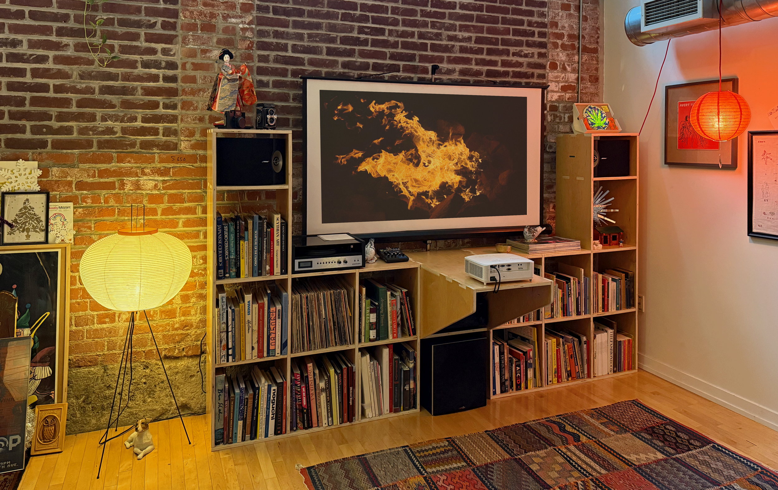 Living room with brick wall, bookshelf, large screen displaying fire, round floor lamp, framed artwork, and a red paper lantern. Wooden floor with patterned rug.