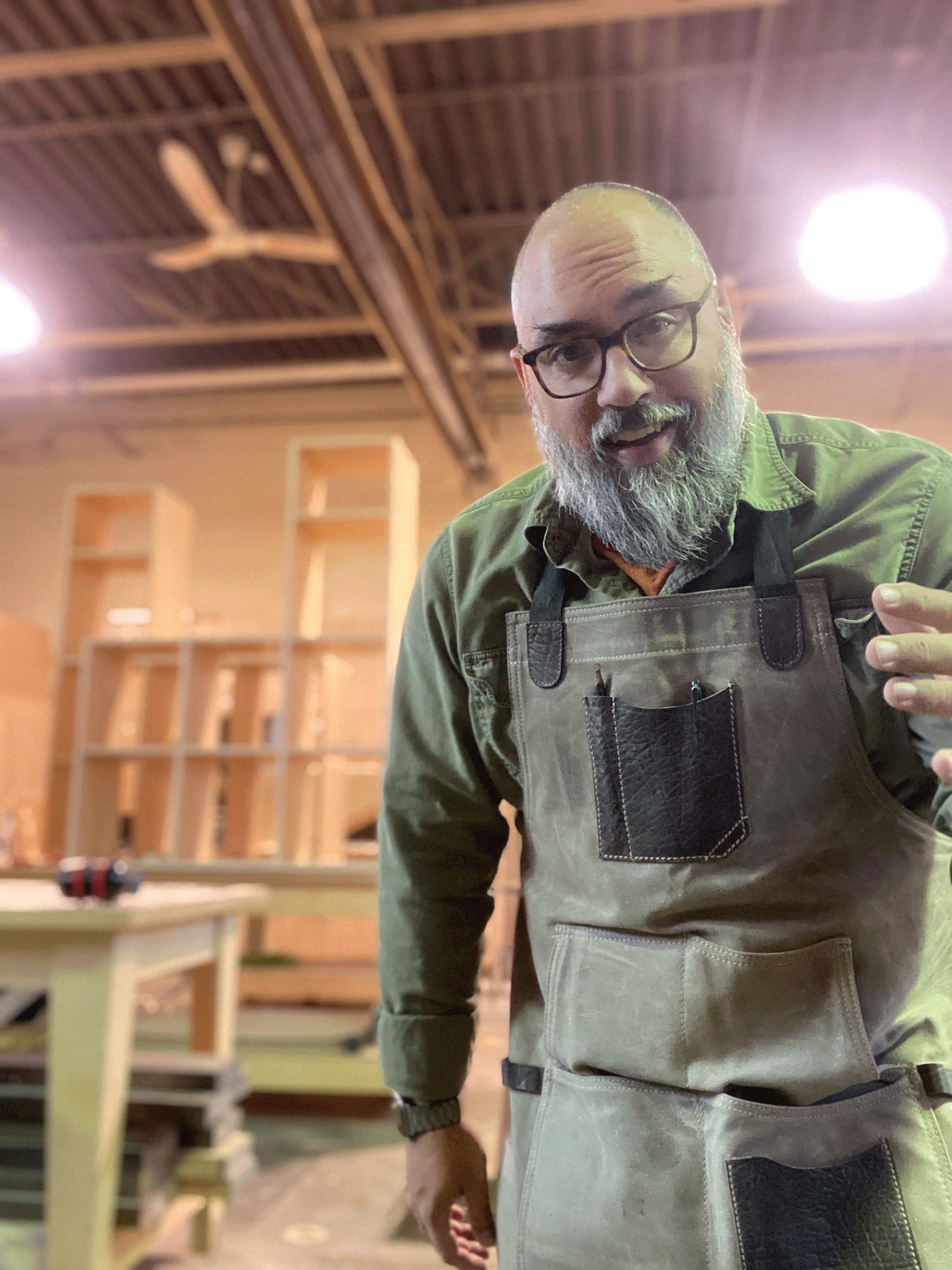A man with glasses and a beard wearing a green shirt and a beige apron, posing in a woodworking shop with unfinished wooden shelves and a table in the background.