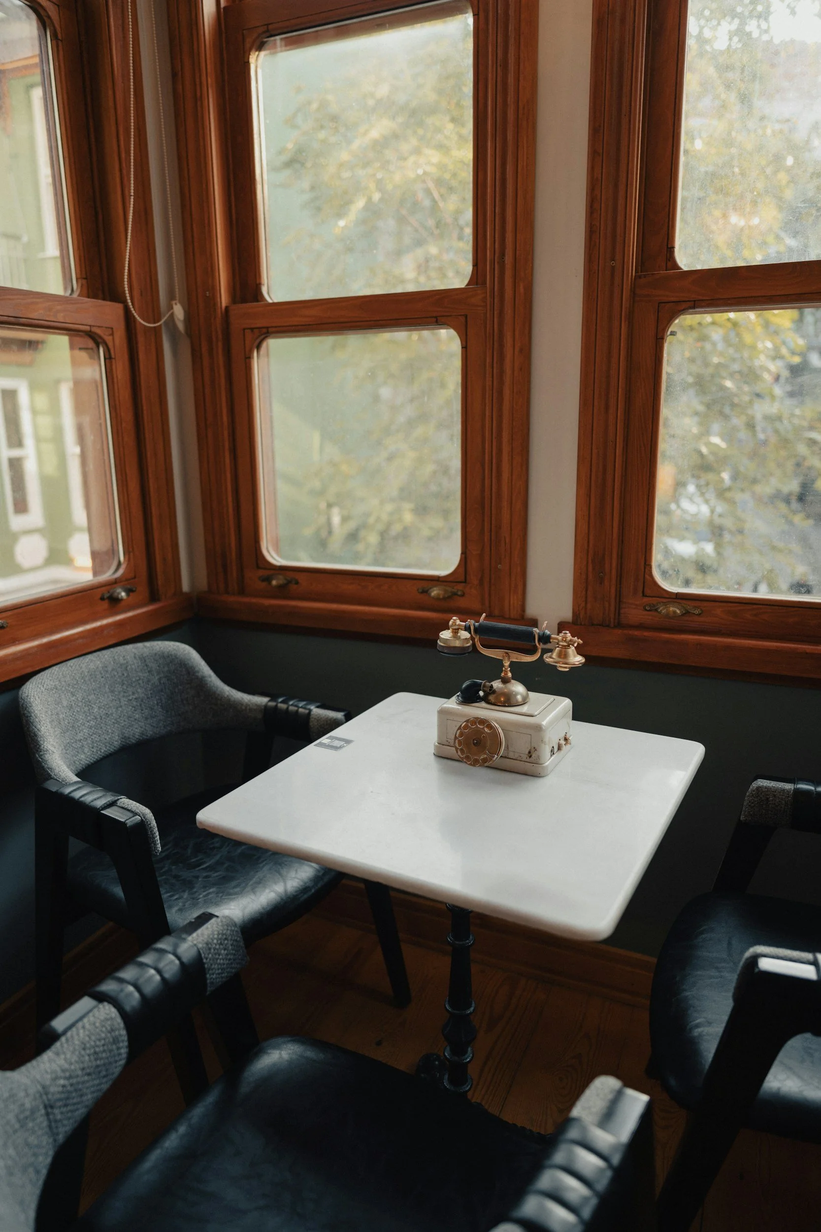 A small white table with a vintage black and brass telephone receiver on top, surrounded by four gray and black armchairs in a room with large wooden-framed windows showing an outdoor scene with trees.