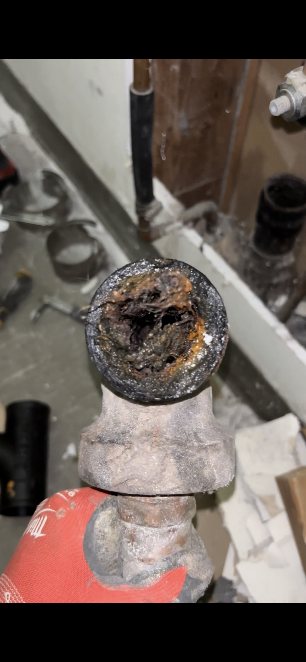 Close-up of rusted and corroded pipe with internal damage, held by a person wearing a red glove in a construction or plumbing setting.