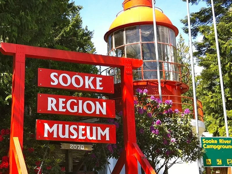 The Sooke Region Museum British Columbia, Canada.  is marked by a large red sign with white letters, situated in front of an orange lighthouse with glass windows and surrounded by trees and purple flowers.