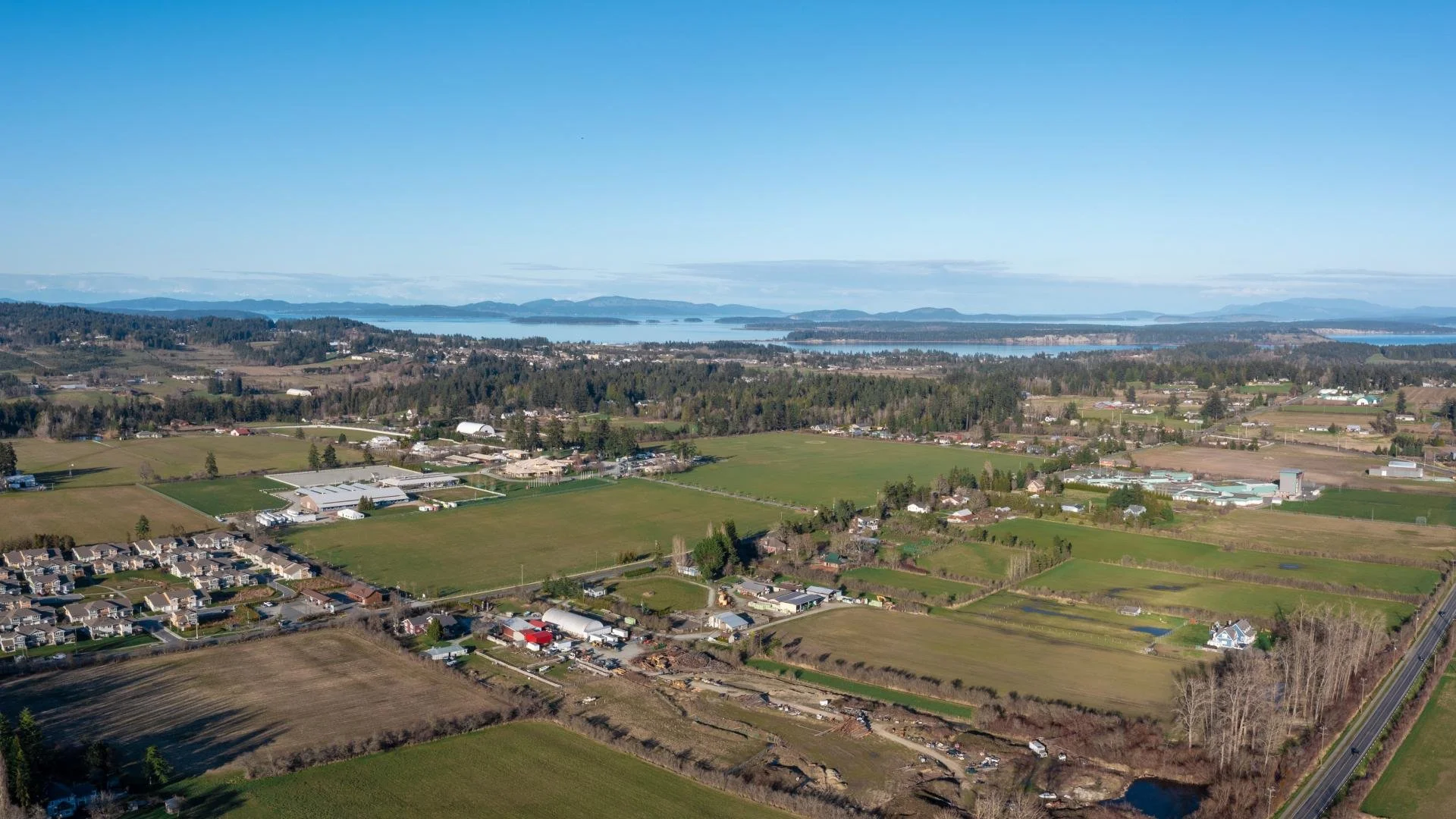 Aerial view of a rural landscape in Central Saanich British Columbia, Canada. with green fields, residential houses, and distant water body mountains.