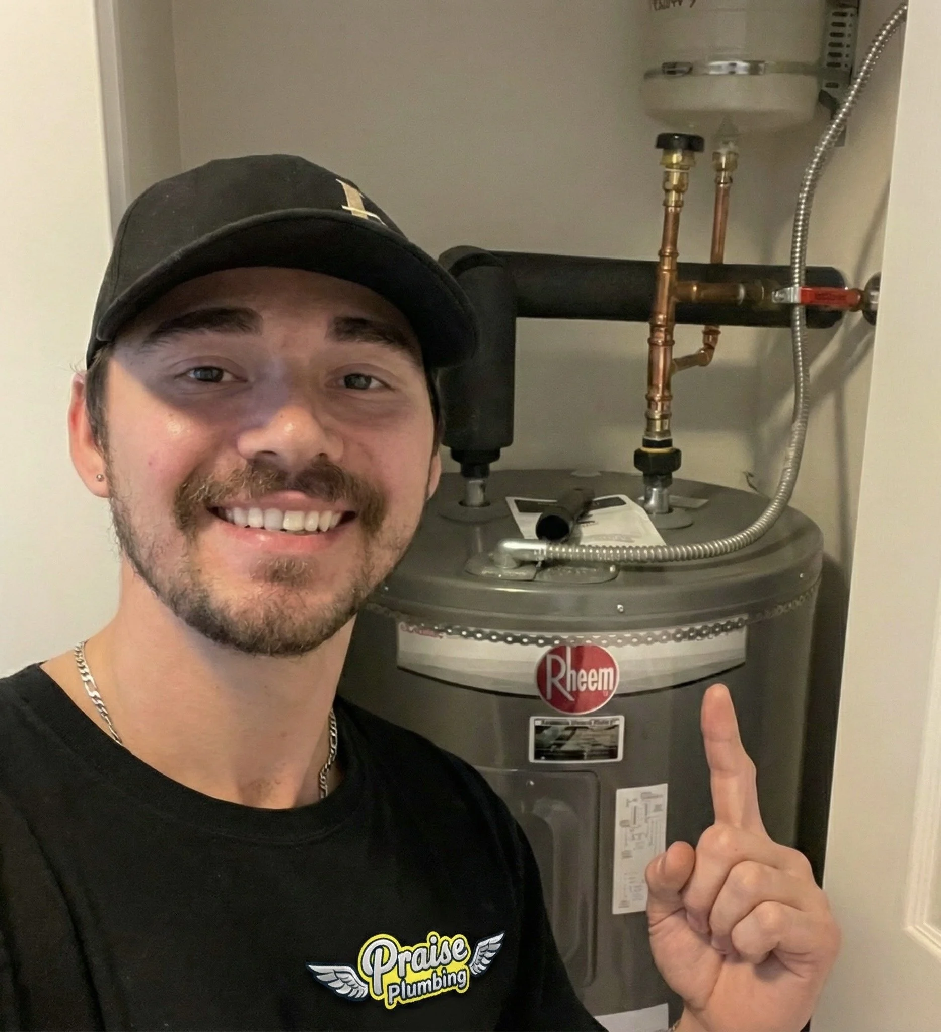 A smiling man wearing a Praise Plumbing t-shirt and a black cap pointing at a Rheem water heater.