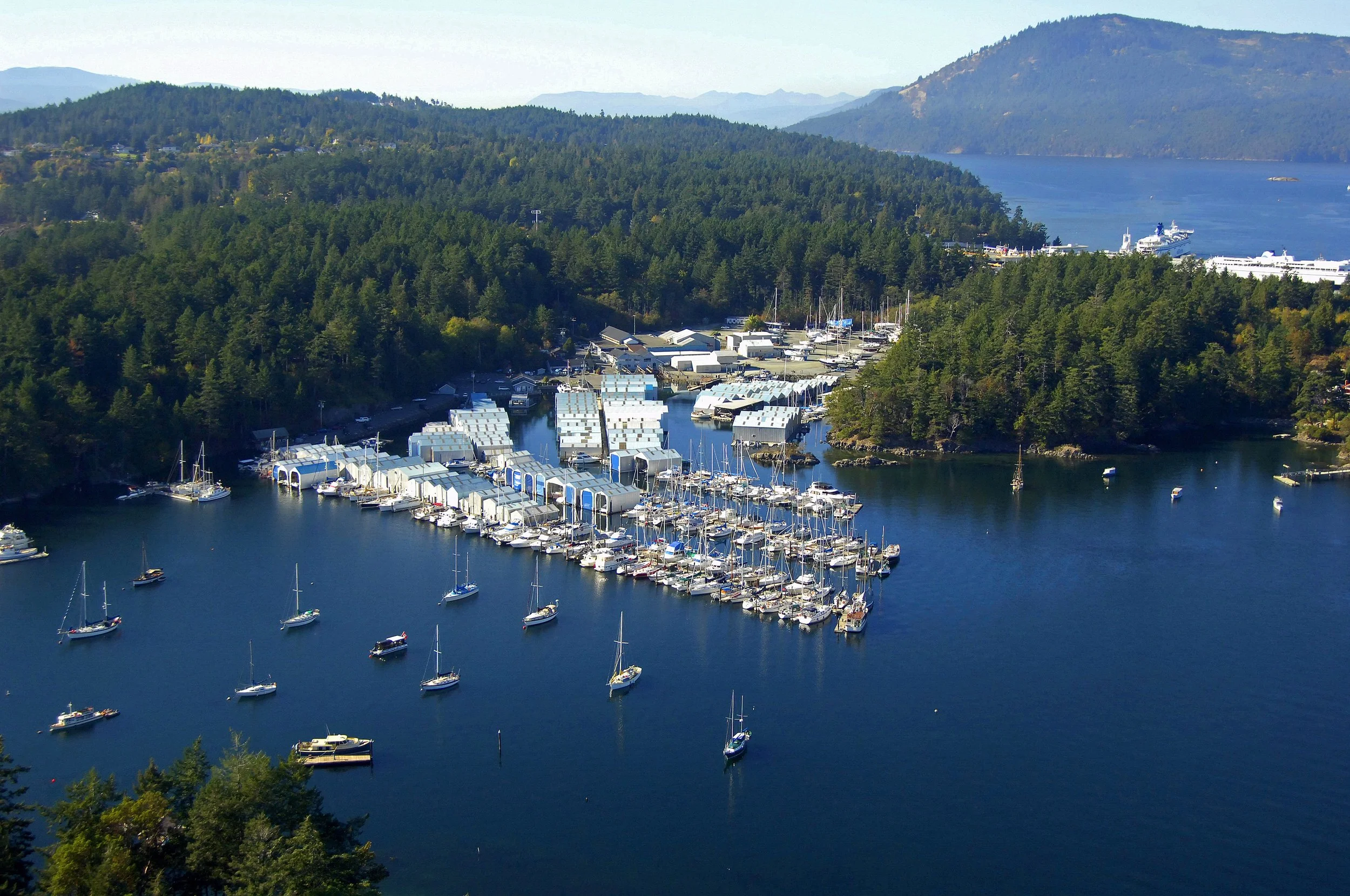 Aerial view of a marina in North Saanich British Columbia, Canada. With numerous sailboats and yachts docked, surrounded by green forests and distant mountains, with a large body of water and ships in the background.