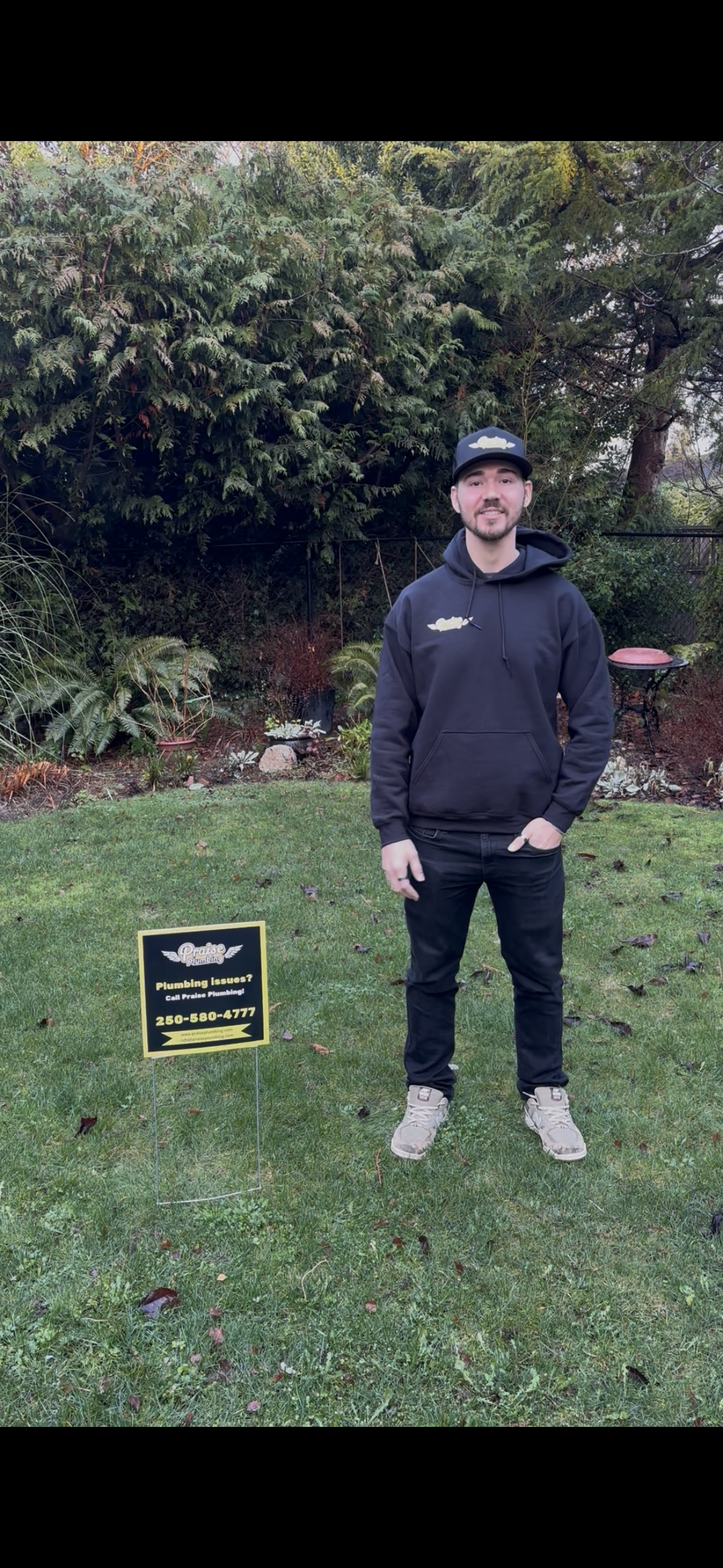 A man standing on a well-maintained lawn in front of a garden with trees and shrubs, wearing a black hoodie and cap, holding a phone in his right hand, with a yard sign advertising plumbing services beside him.