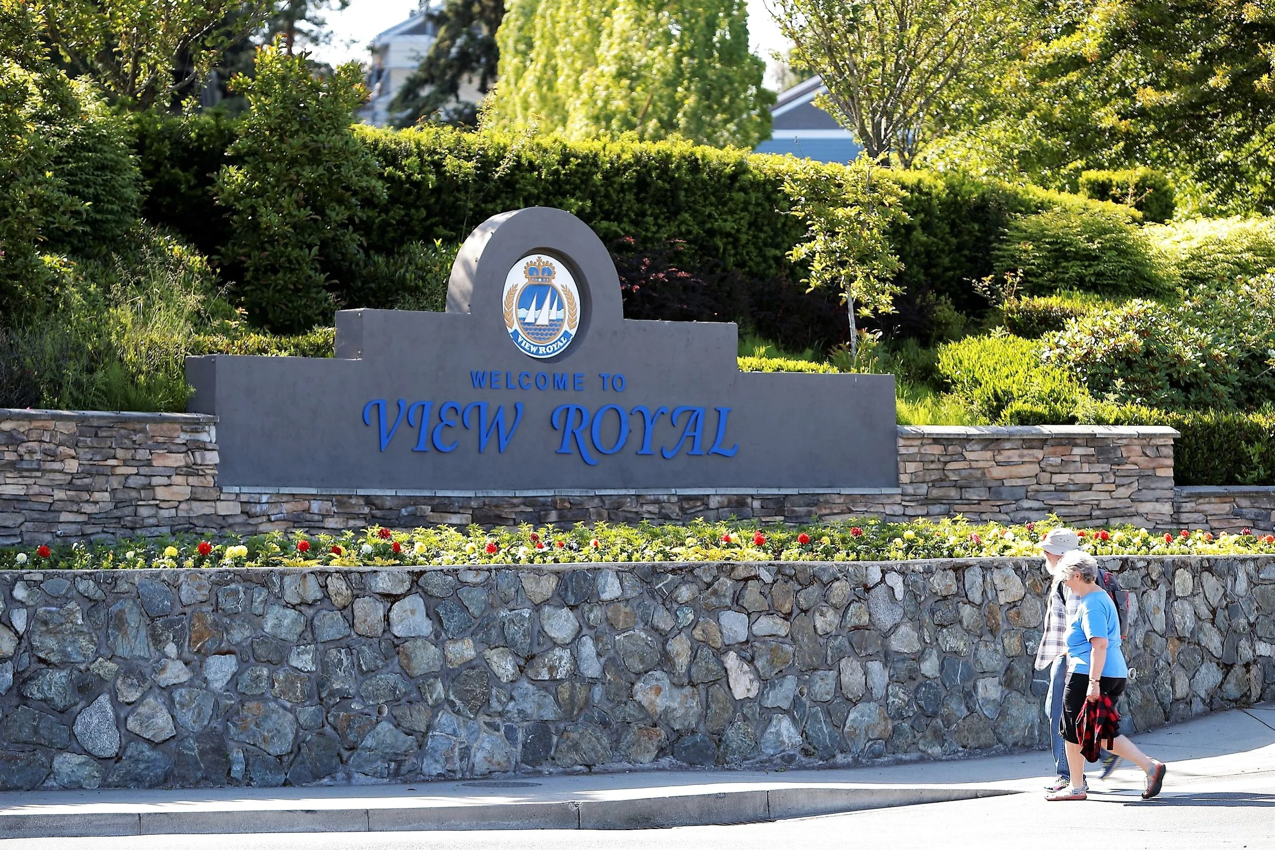 Sign that reads 'Welcome to View Royal' with a crest above, surrounded by greenery and a stone wall, and two people walking in front of it.