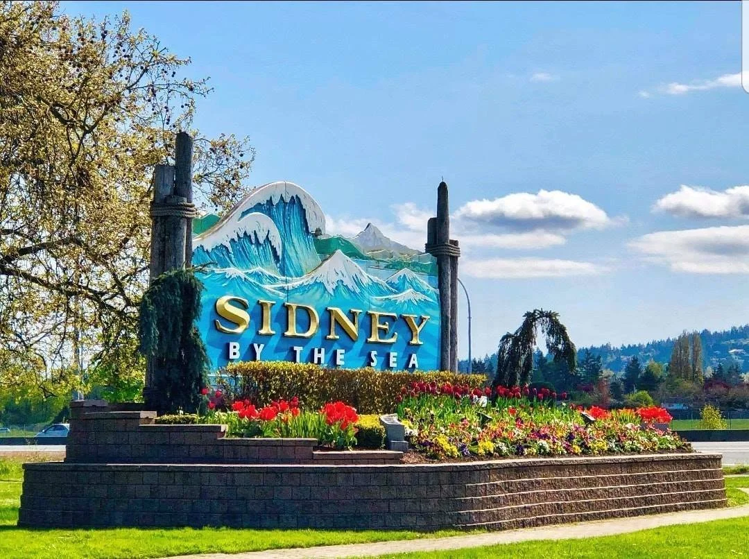 Sign reading "Sidney by the Sea" with a colorful painted mountain and wave design, surrounded by flower beds and trees under a blue sky with some clouds.