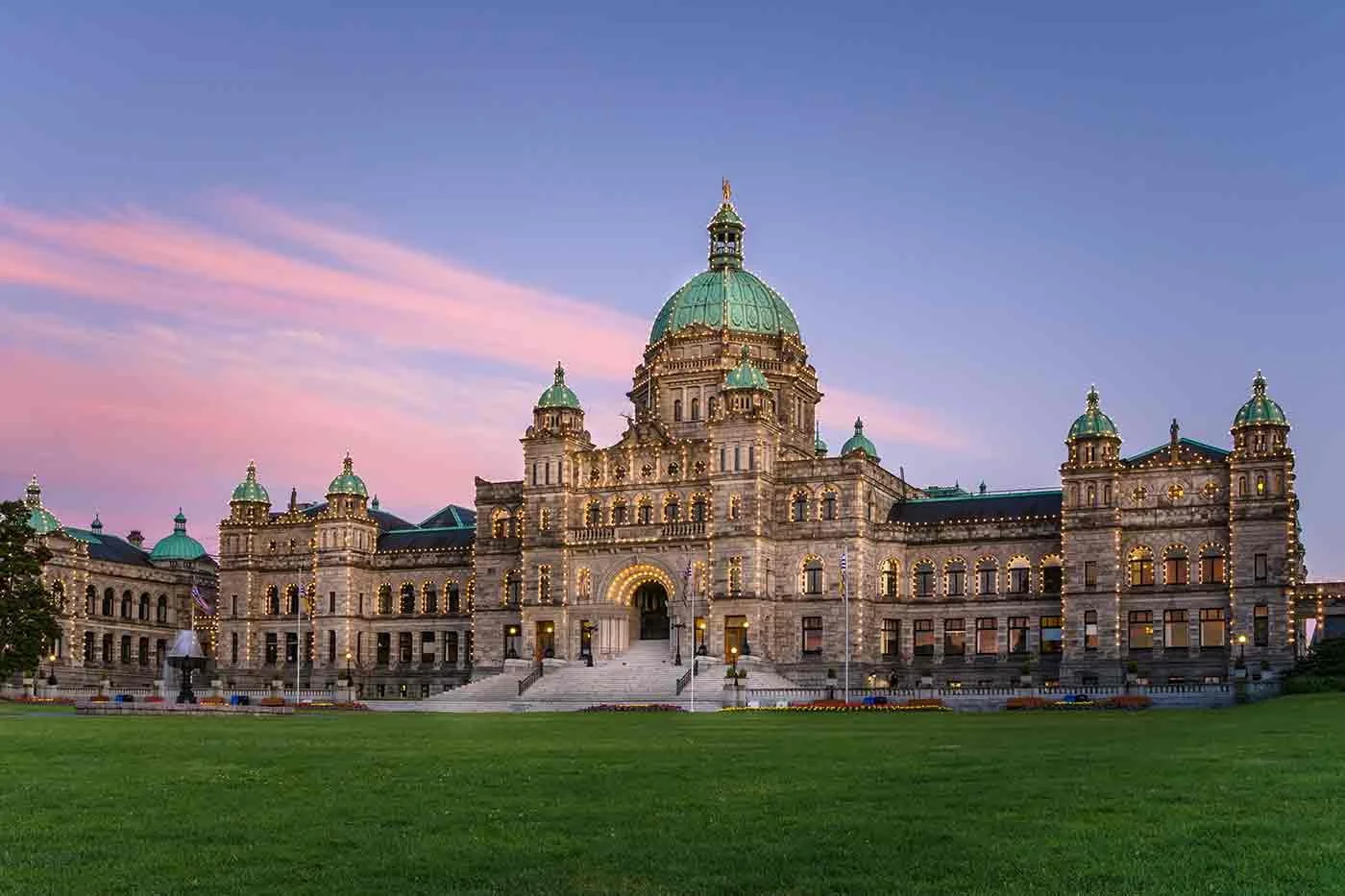 The British Columbia Parliament Buildings in Victoria, Canada, illuminated at dusk with a clear sky and pink clouds.