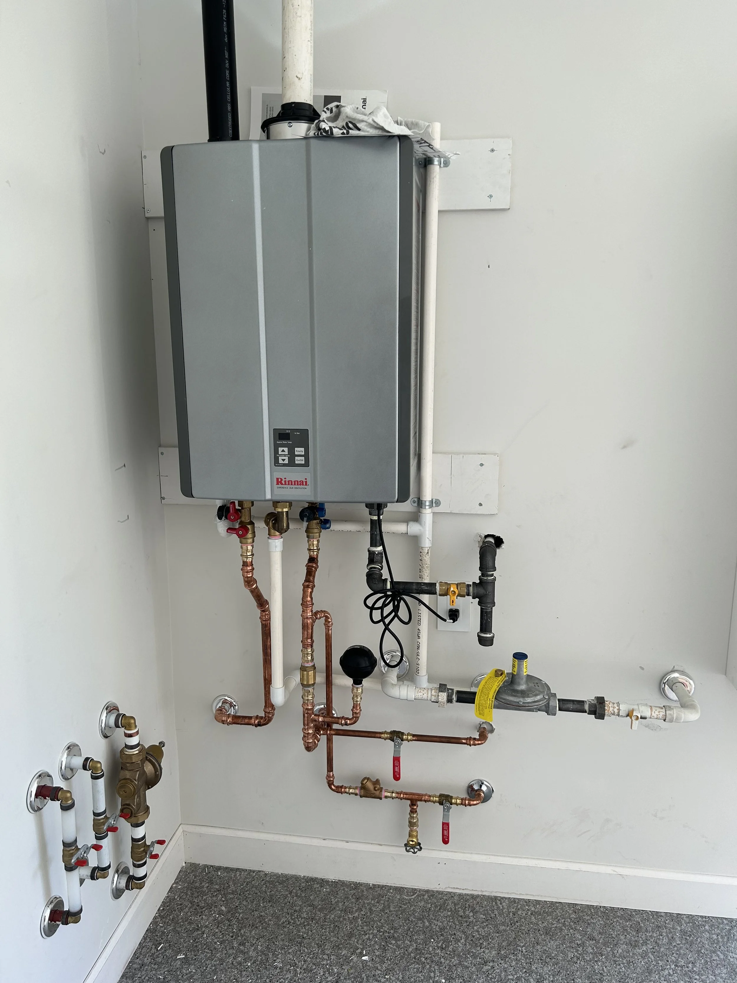 Installation of a tankless water heater with copper, plastic, and black pipes connected to the unit on a white wall, with red and blue valves and a water meter below.