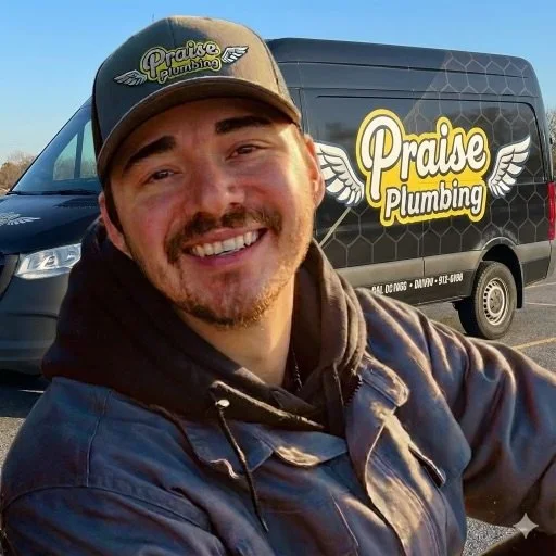 A man smiling in front of a black service van with a 'Praise Plumbing' logo. The man is wearing a baseball cap and a jacket.
