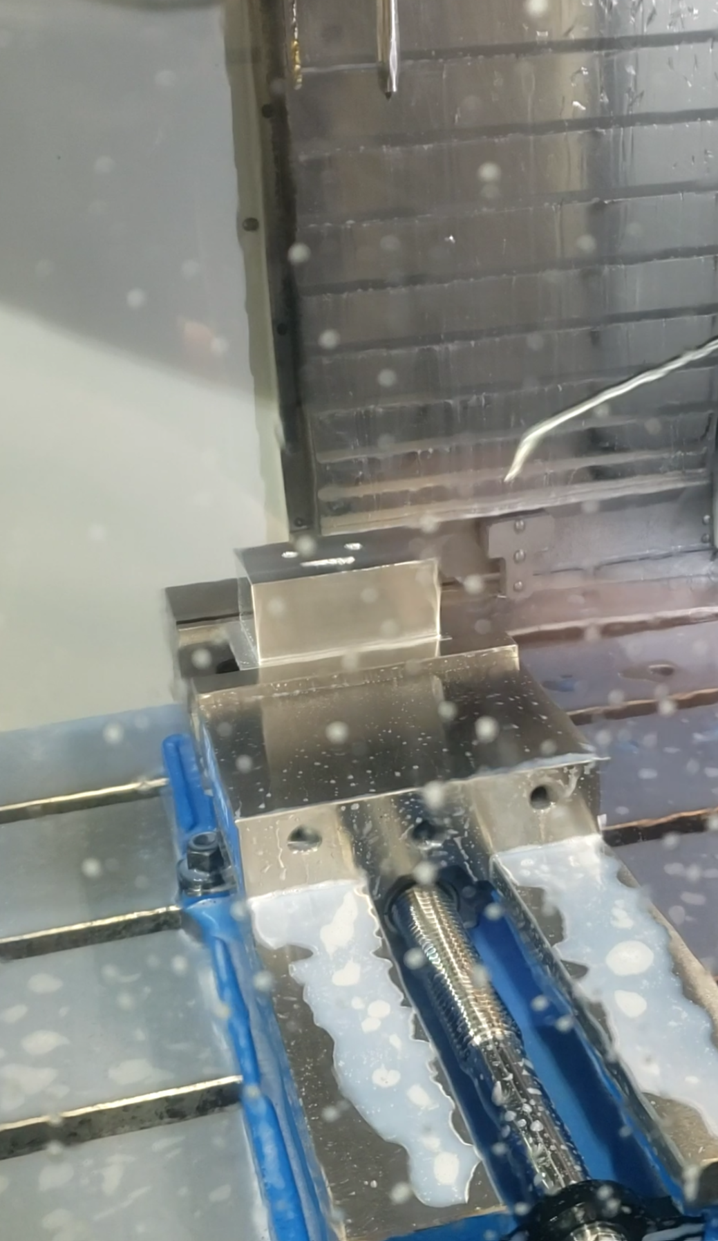 Close-up of a metal CNC machine part, with coolant or lubricant splattered on its surface, and a rotating drill bit or spindle.