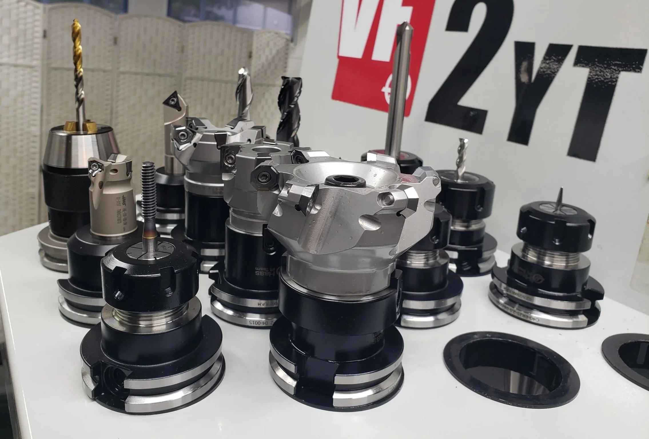 Various industrial machine parts and tools, including drill bits and other precision components, arranged on a white surface at an exhibition or workshop.