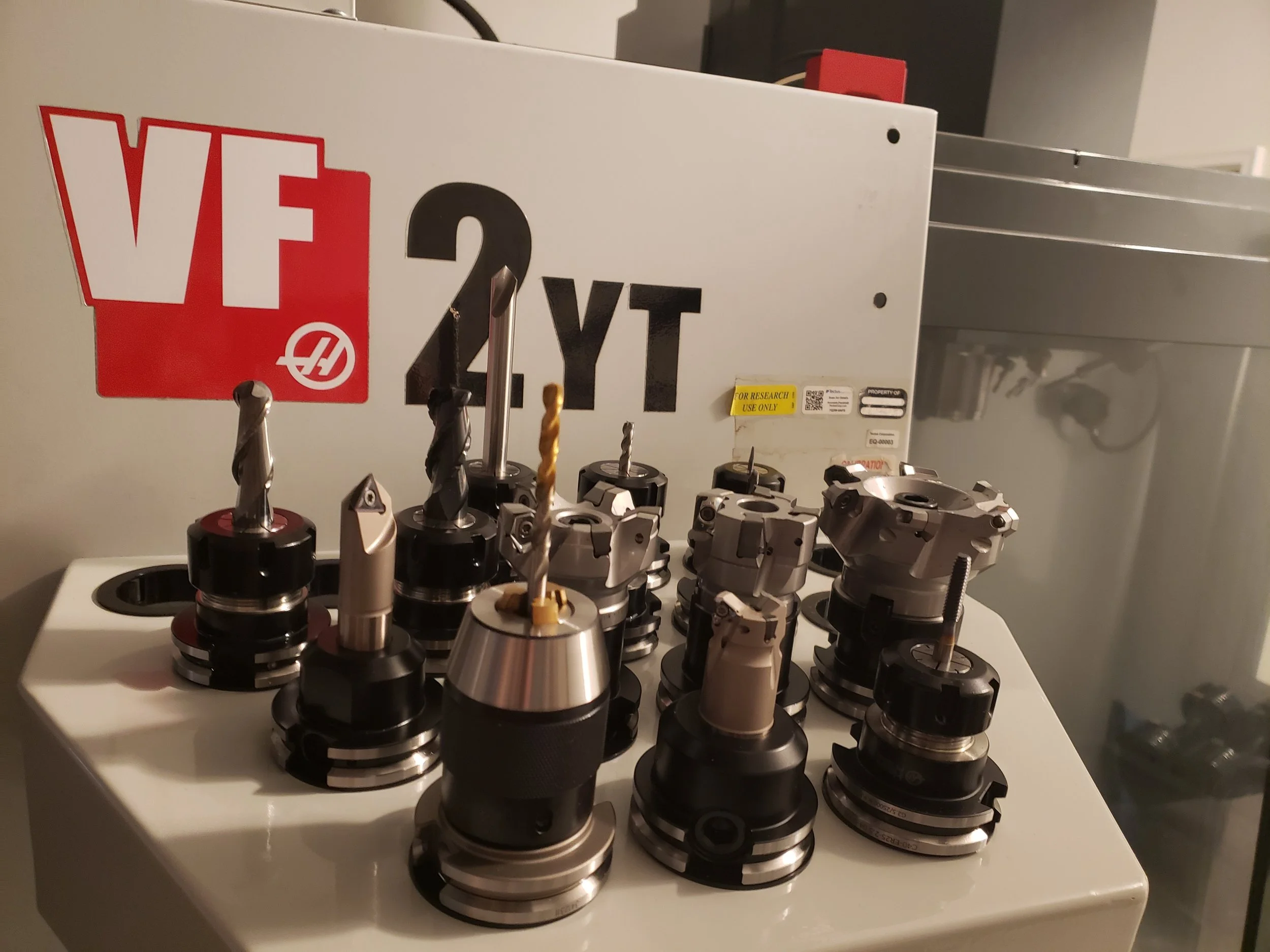 Collection of various CNC milling machine tools and drill bits arranged on a white surface, with a machine label in the background.