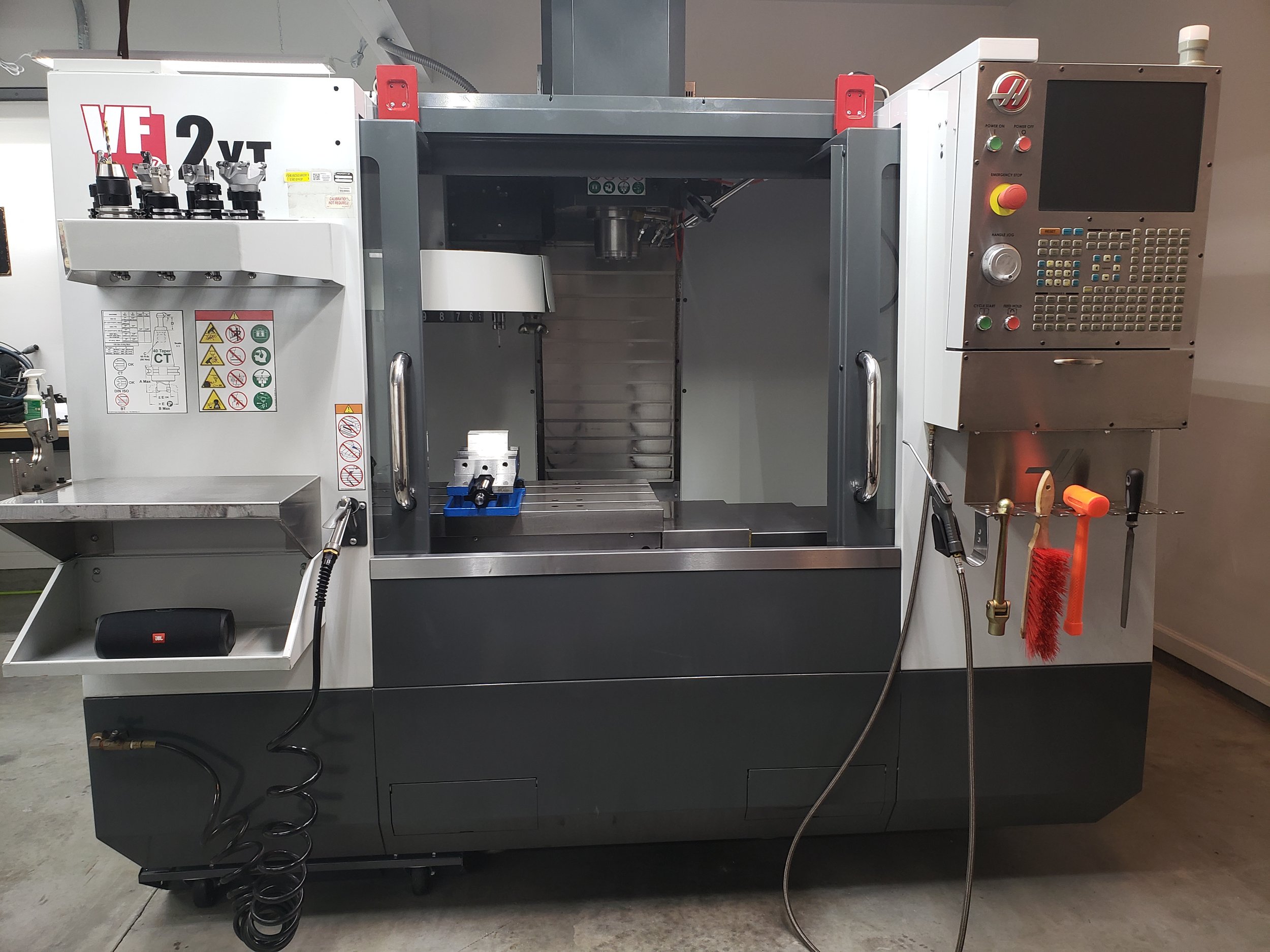 A large CNC machine with a computer control panel on the right side, various tools hanging below, and safety and operation labels on the front. The machine is in a workshop setting.