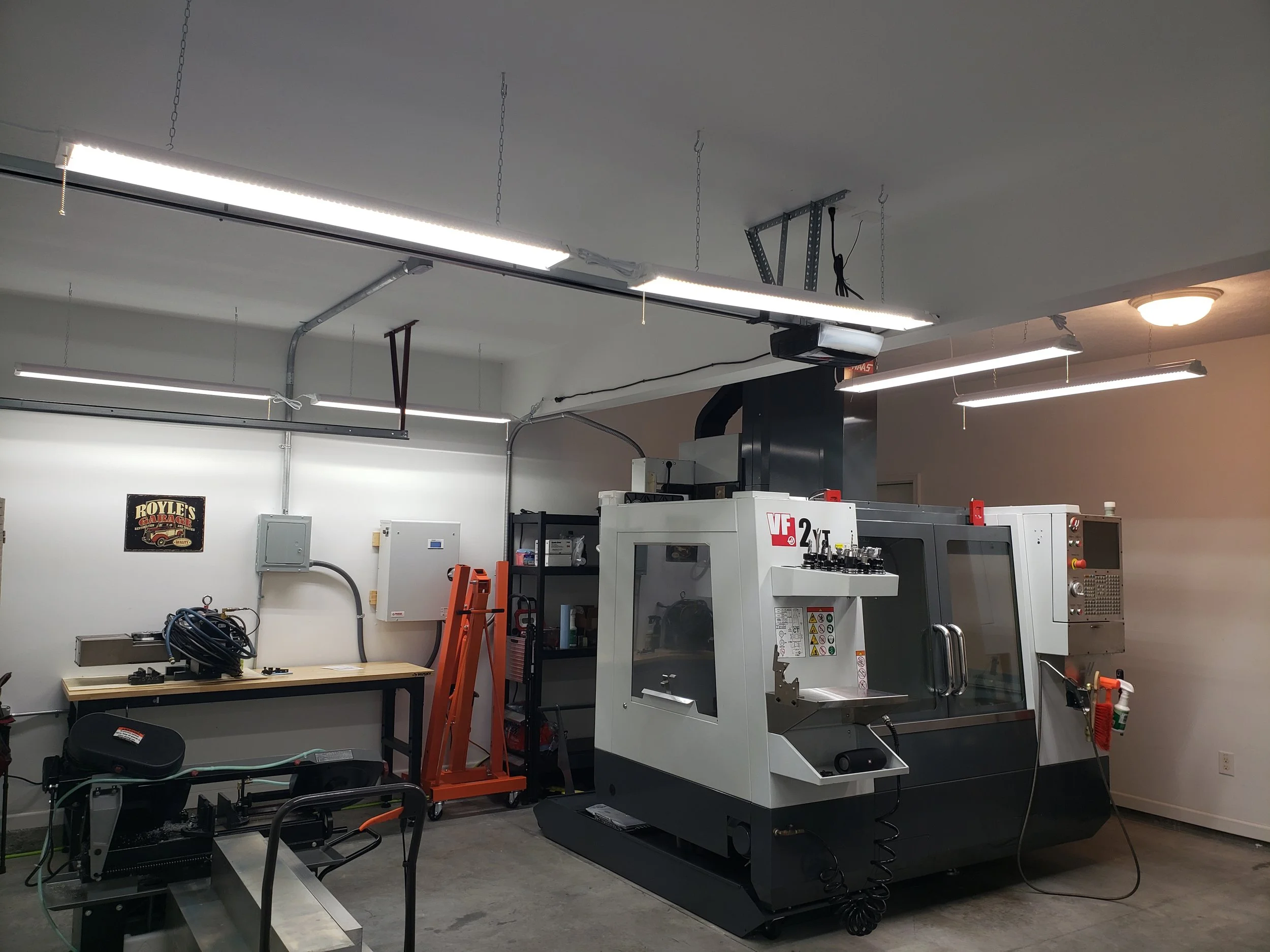 A workshop contains a large industrial machine, a workbench with tools, and various equipment including a foldable ladder, a toolbox, and electrical panels. The room has overhead lighting and a plain wall with a sign.