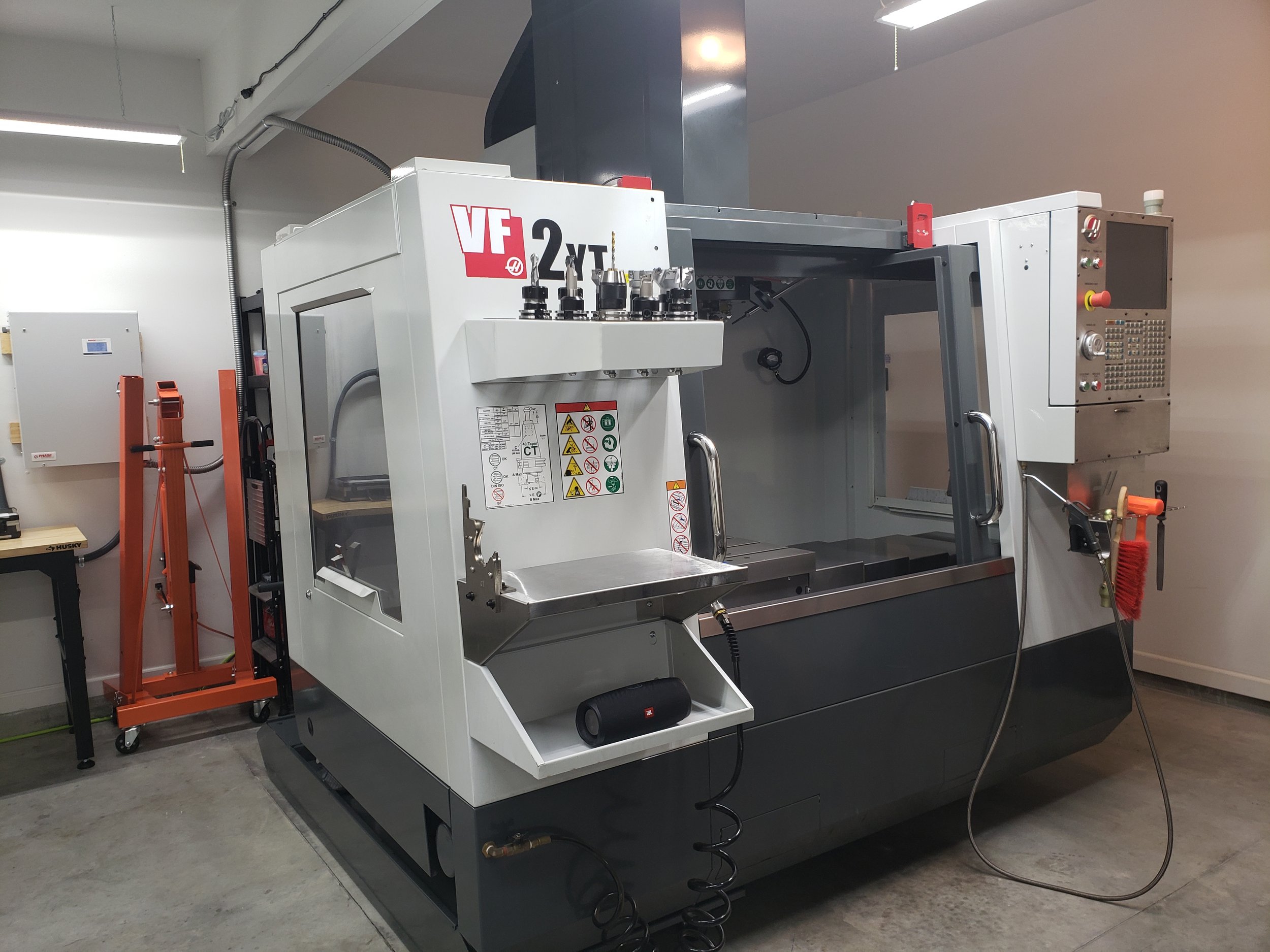 A CNC machine with a control panel on the right side, safety warnings, and a transparent door, situated in a workshop with a concrete floor and white walls.