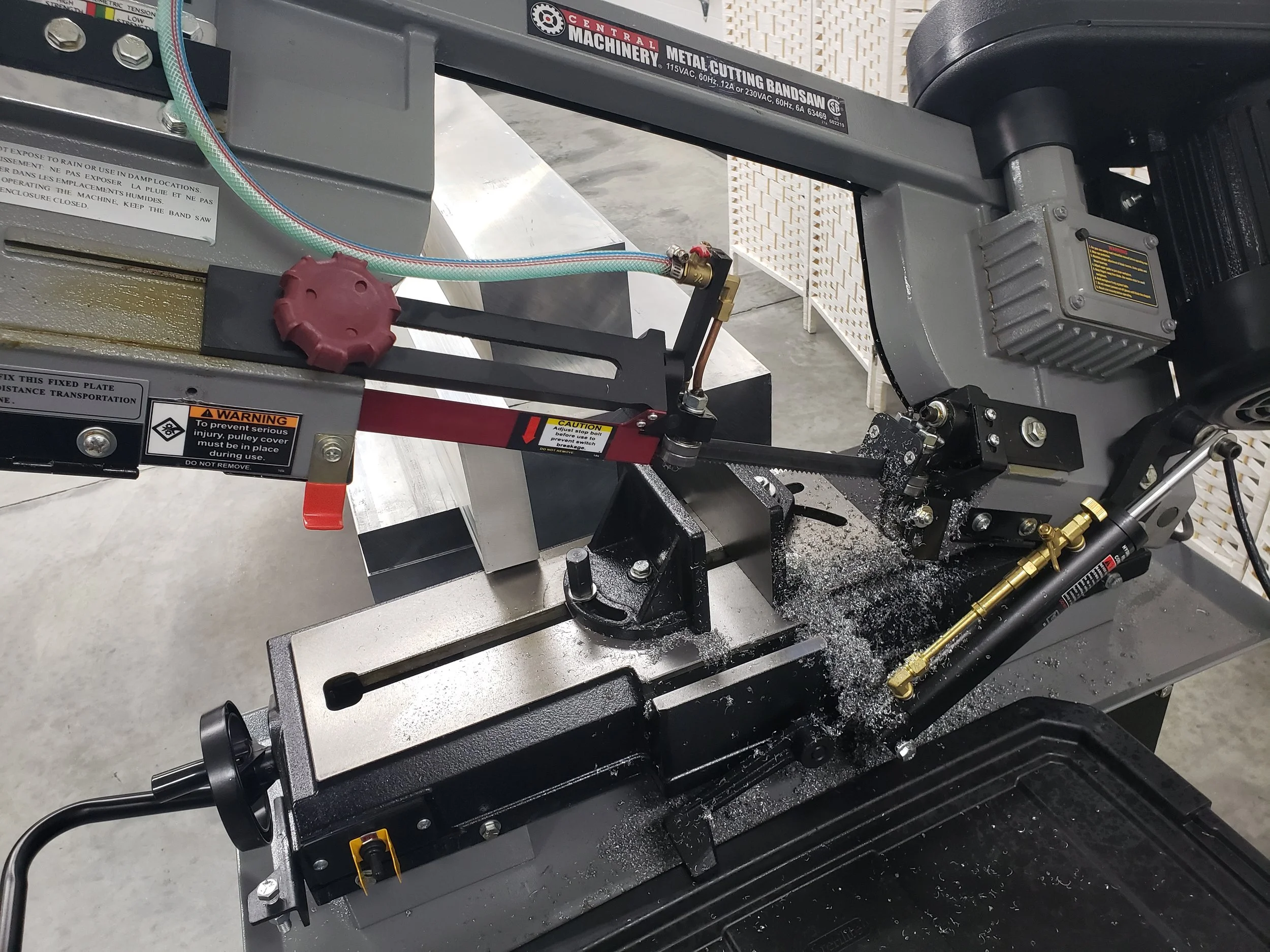 A metal band saw machine in a workshop, with metal shavings and liquid coolant on its black work surface, warning and caution labels, and a motor on the right side.