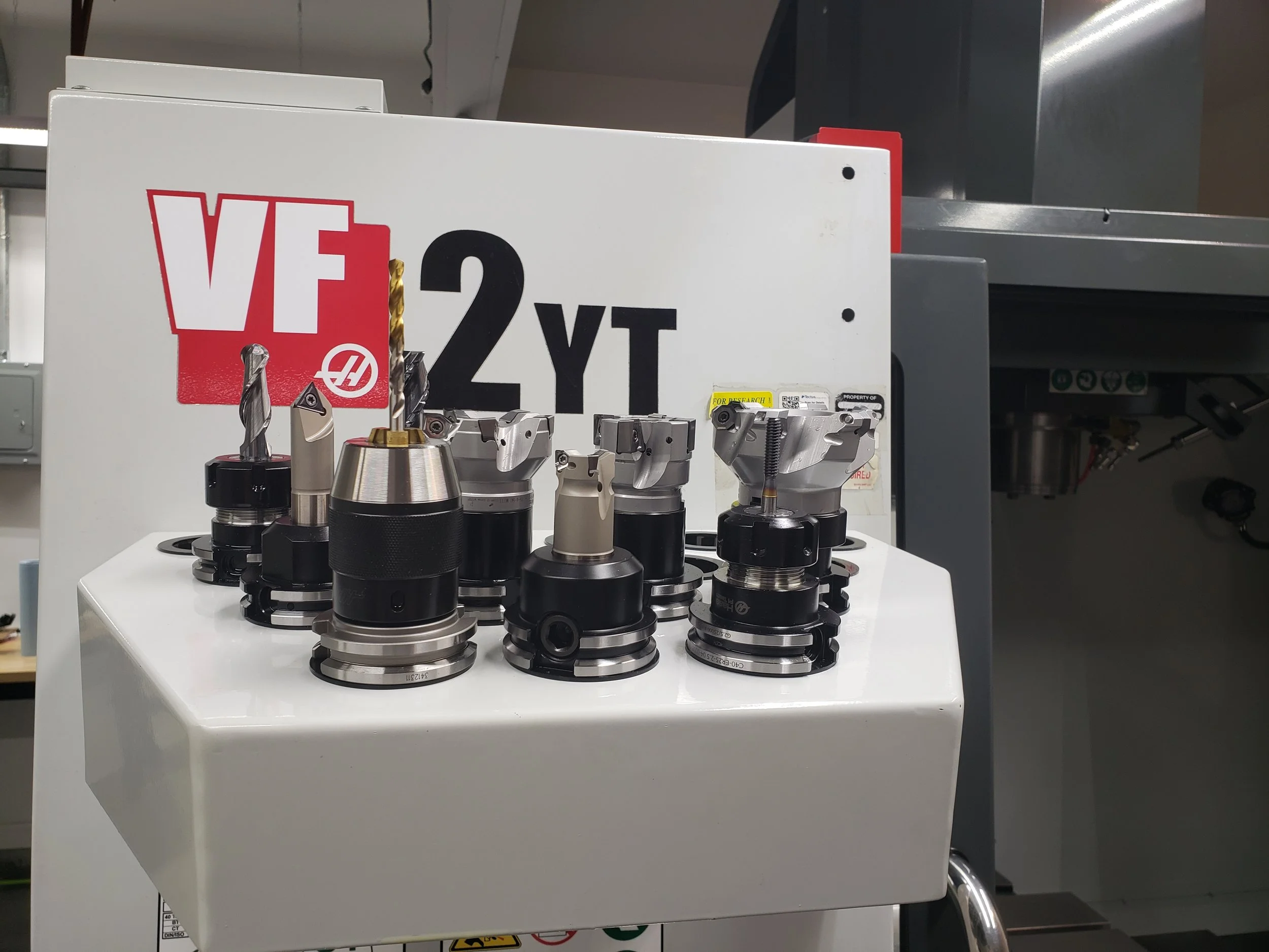 Various CNC machine tool holders displayed on a white stand in a manufacturing environment, with a partially visible programming sign in the background.