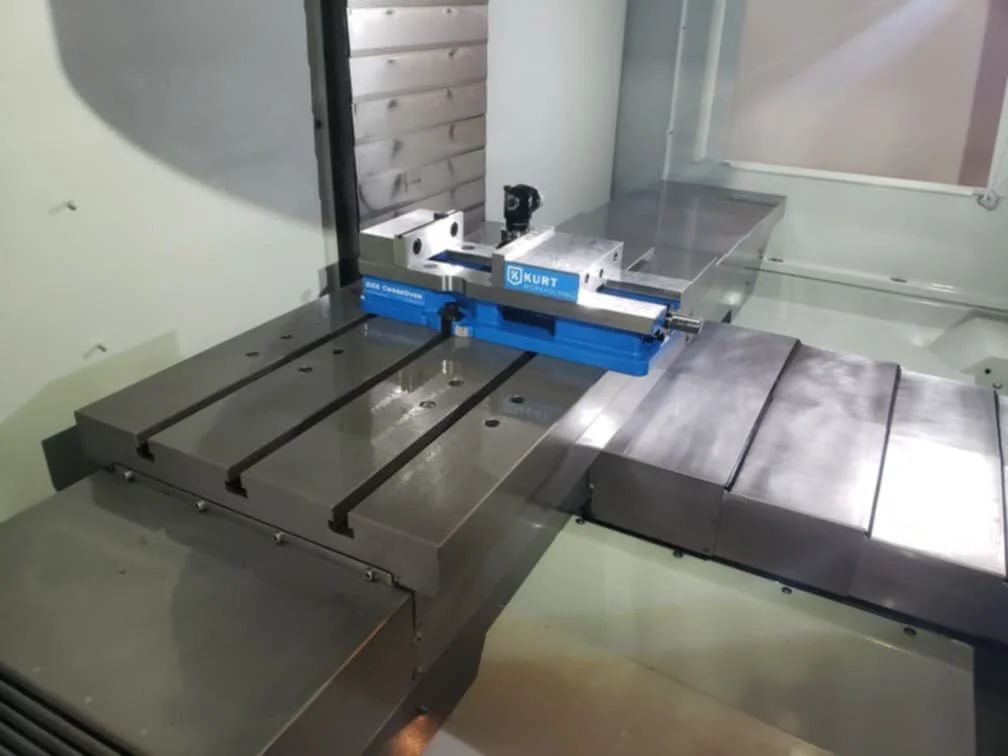 Close-up of a precision machining or manufacturing machine station with metallic components and a blue fixture marked 'KURT'.