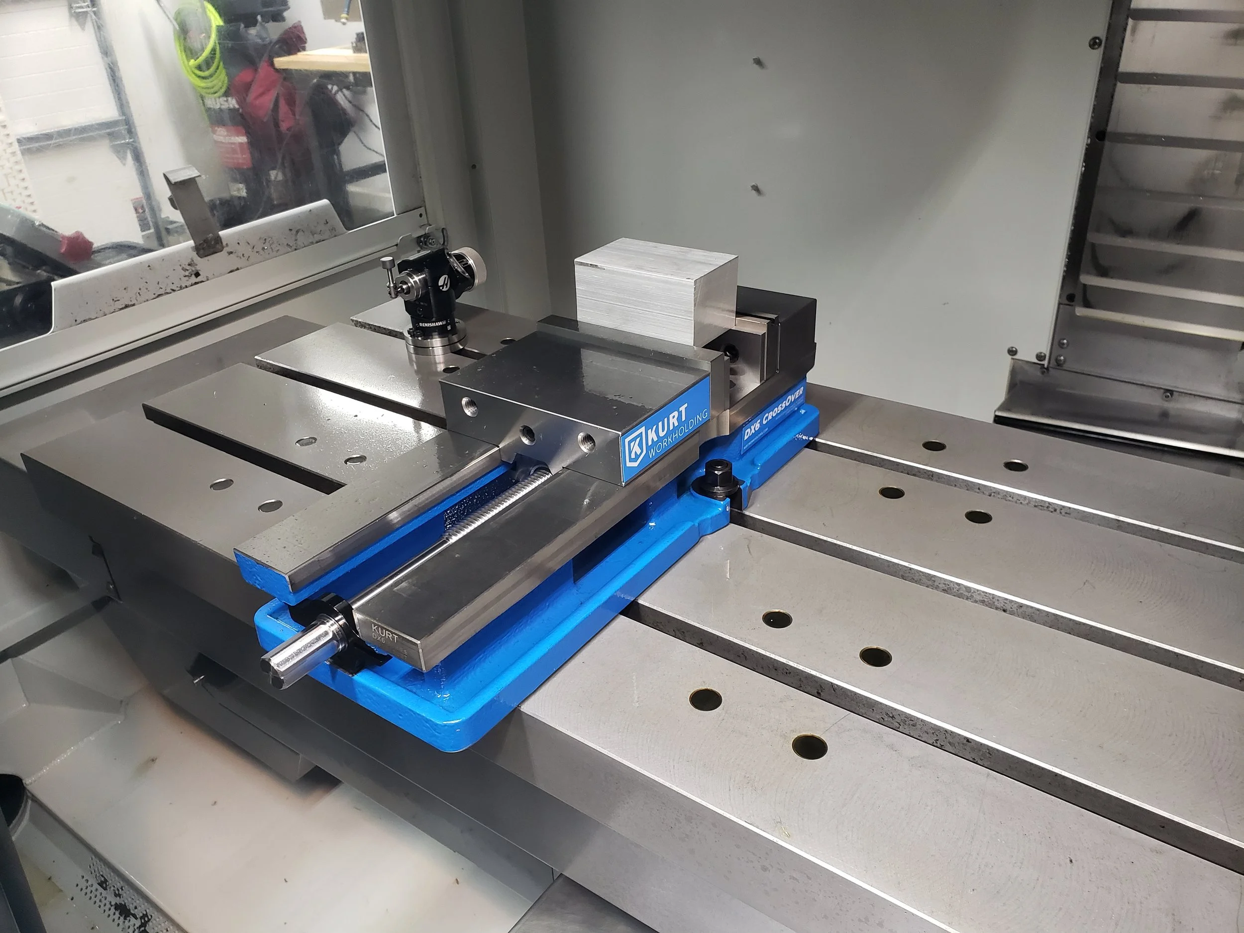 A milling machine with a silver block clamped on the work surface, setup for machining, with the machine and surrounding workspace visible.