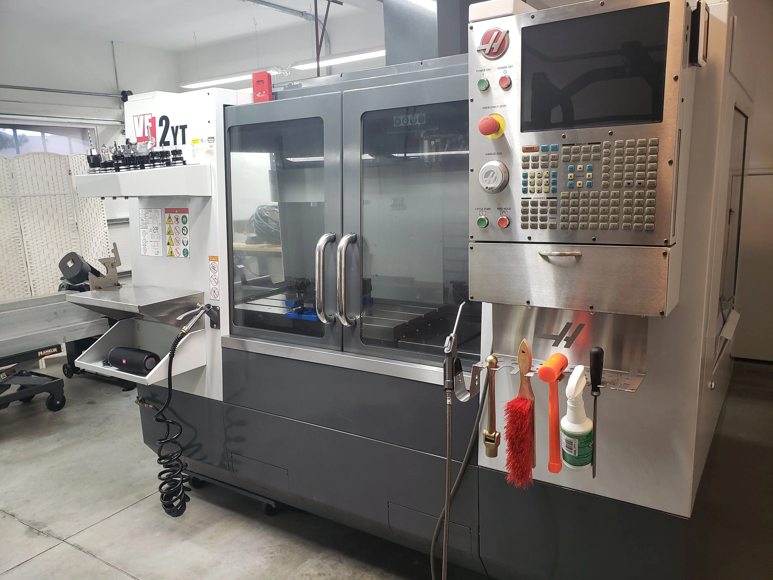 Industrial CNC machine with control panel, protective glass doors, and various tools hanging on the machine.