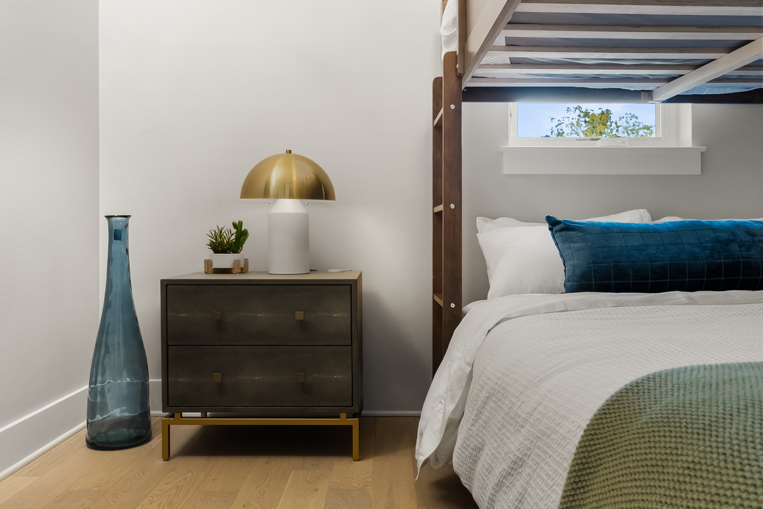 A bedroom corner with a bed, a nightstand, a lamp, a vase, and a small potted plant. A window is above the bed showing trees outside.