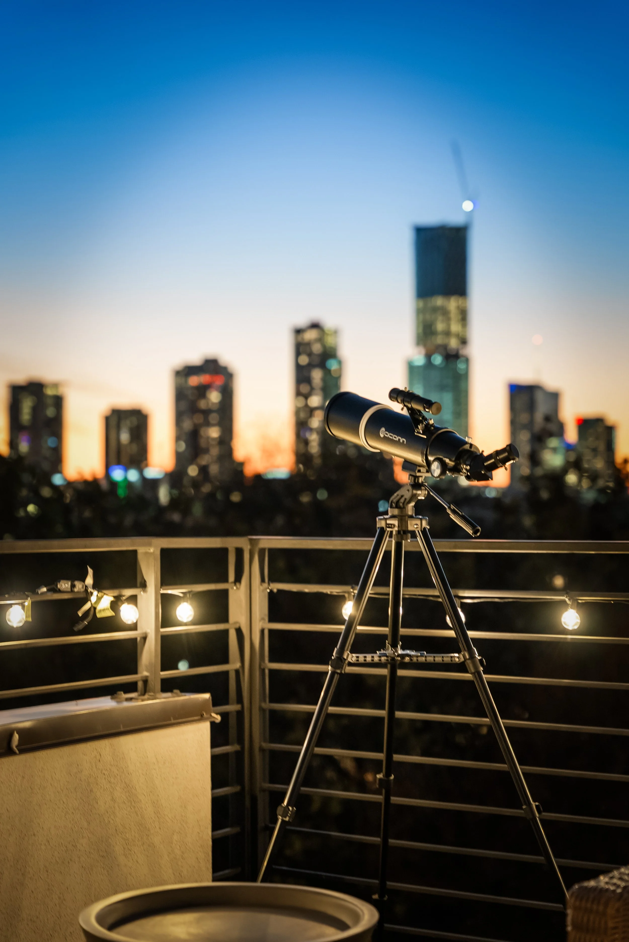 A telescope set up on a balcony railing with city skyline in the background during sunset.